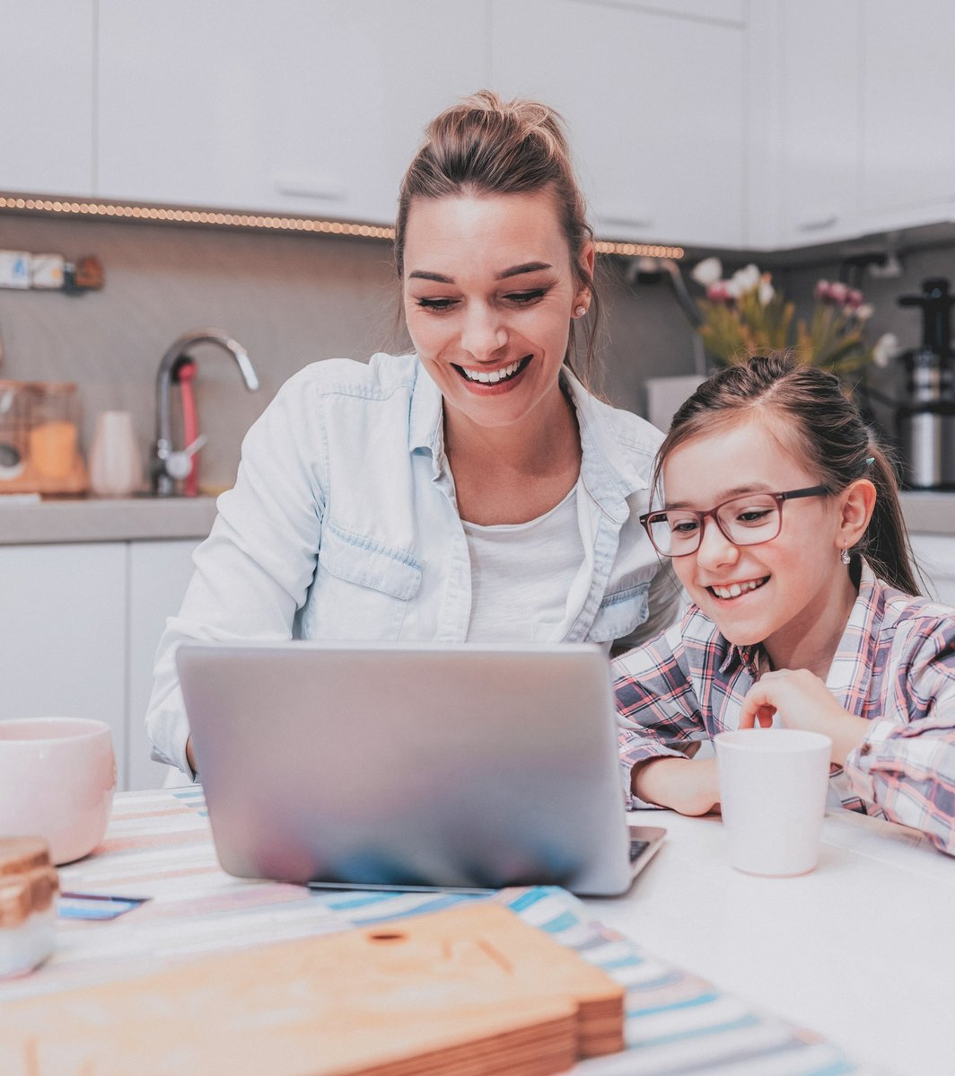 An adult woman and a young girl are happily looking at a laptop screen together at a kitchen table. They both smile as the woman puts her arm around the child, creating a warm, engaged atmosphere.