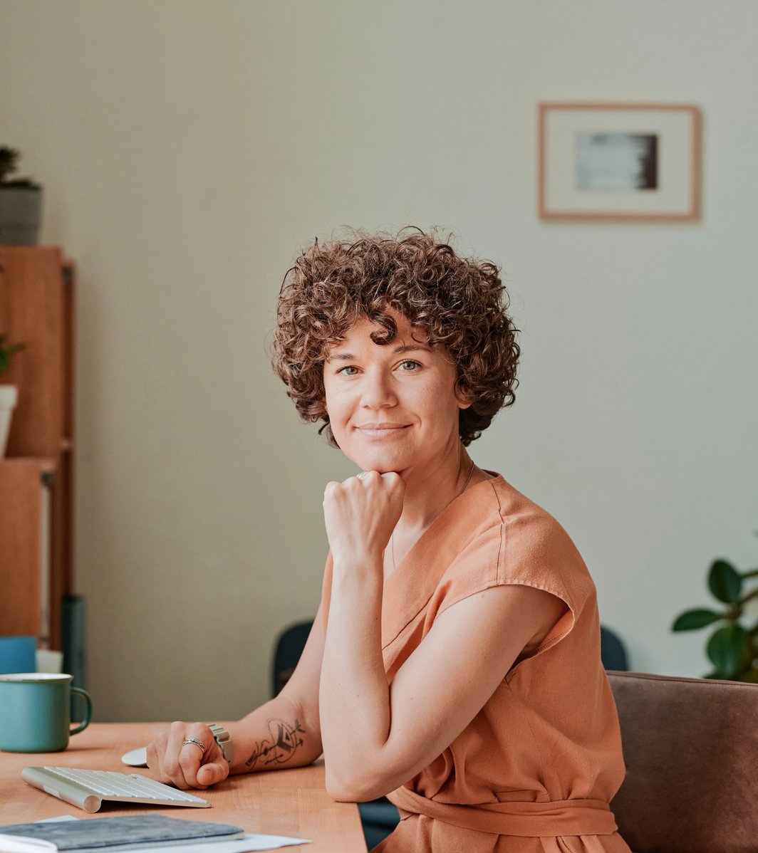 A smiling adult with curly hair sits at a modern desk in an office setting, looking directly at the viewer with a friendly expression. A computer monitor and keyboard are visible on the desk.
