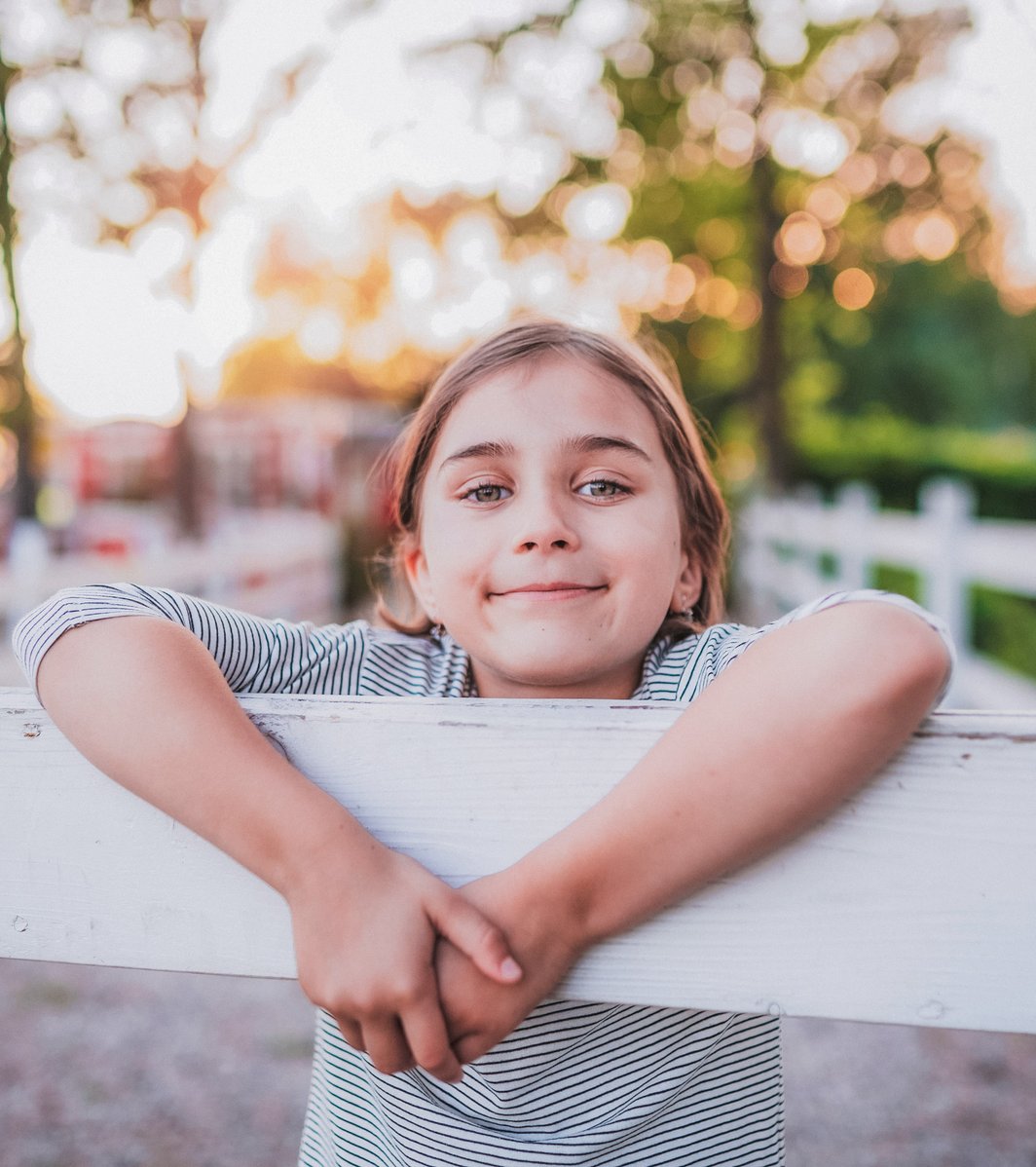 A happy young child leans on a white wooden fence outdoors, smiling gently at the viewer. The background is softly blurred with green trees and warm, bright sunlight.