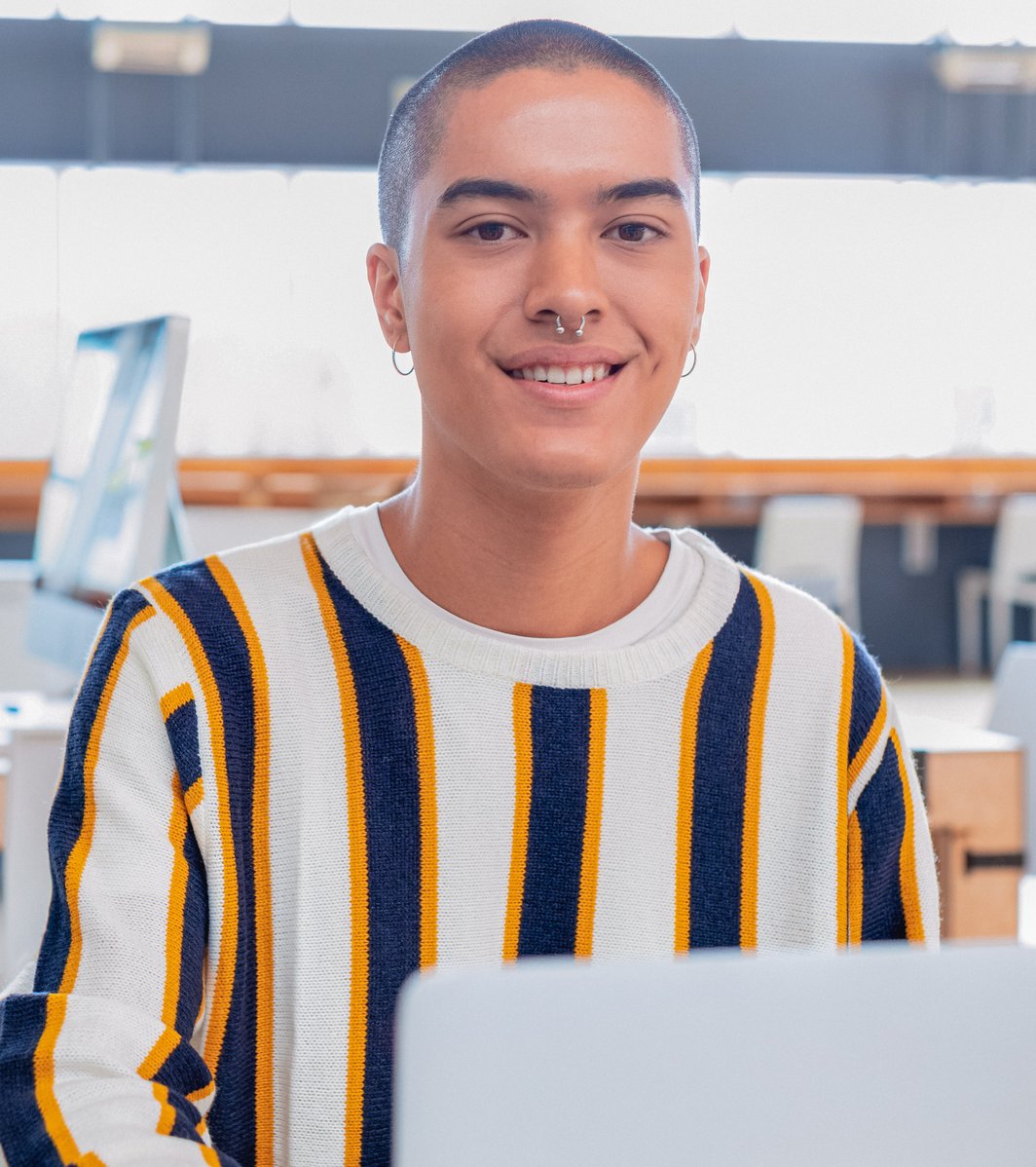 A smiling young adult with a shaved head and nose piercing looks directly at the camera while sitting at a desk with a laptop in a bright, modern indoor setting. They appear friendly and engaged.