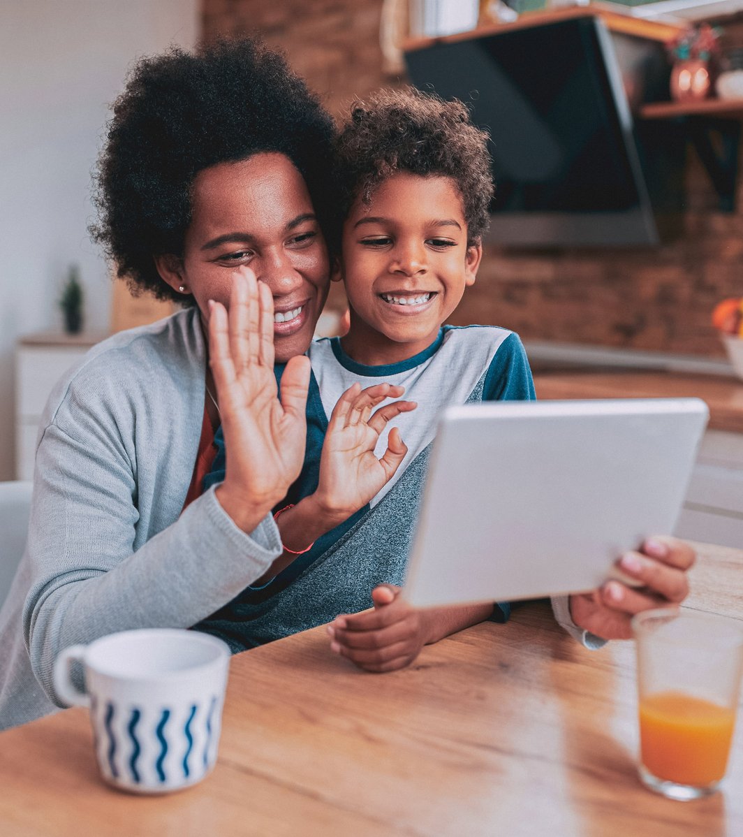 An adult woman and a young boy are smiling and waving while looking at a tablet together in a bright kitchen. They appear happy and engaged in a video call or digital activity.