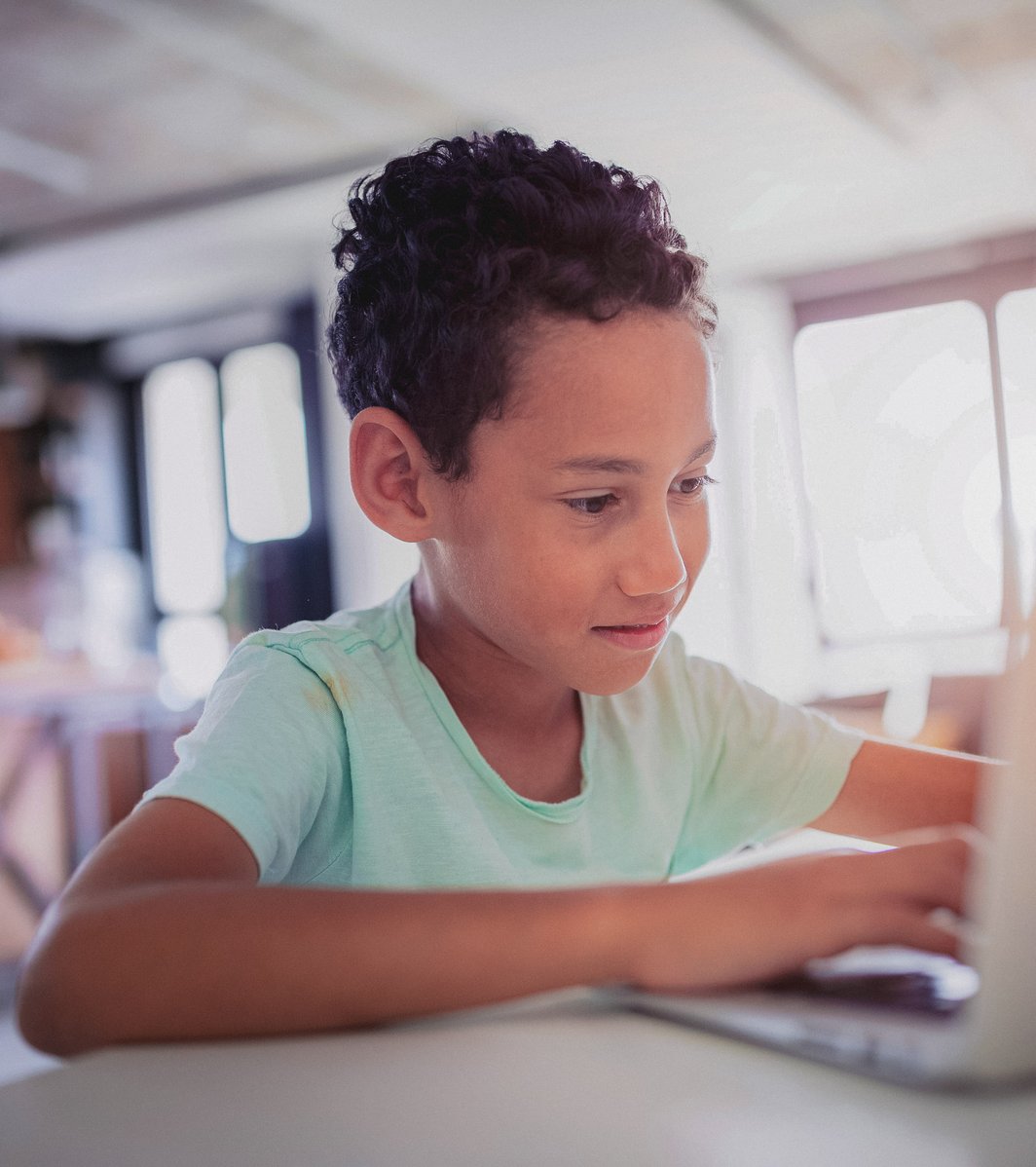 A young boy with curly hair is focused on typing on a laptop, which is placed on a light-colored table in an indoor setting. He appears engaged with the screen.