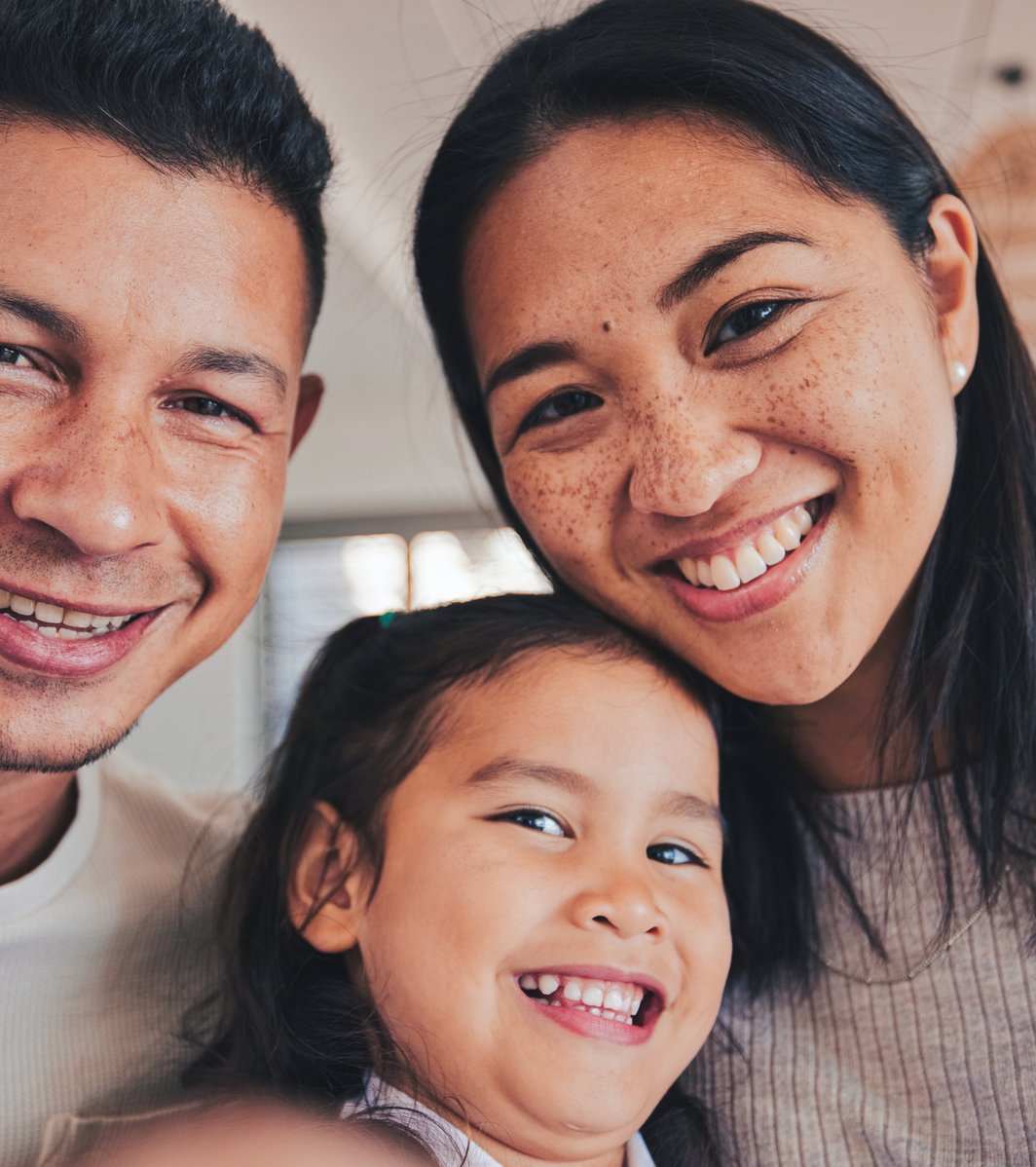 A close-up, smiling portrait of an adult man, an adult woman, and a young child taken indoors, conveying a sense of warmth and happiness among the family.