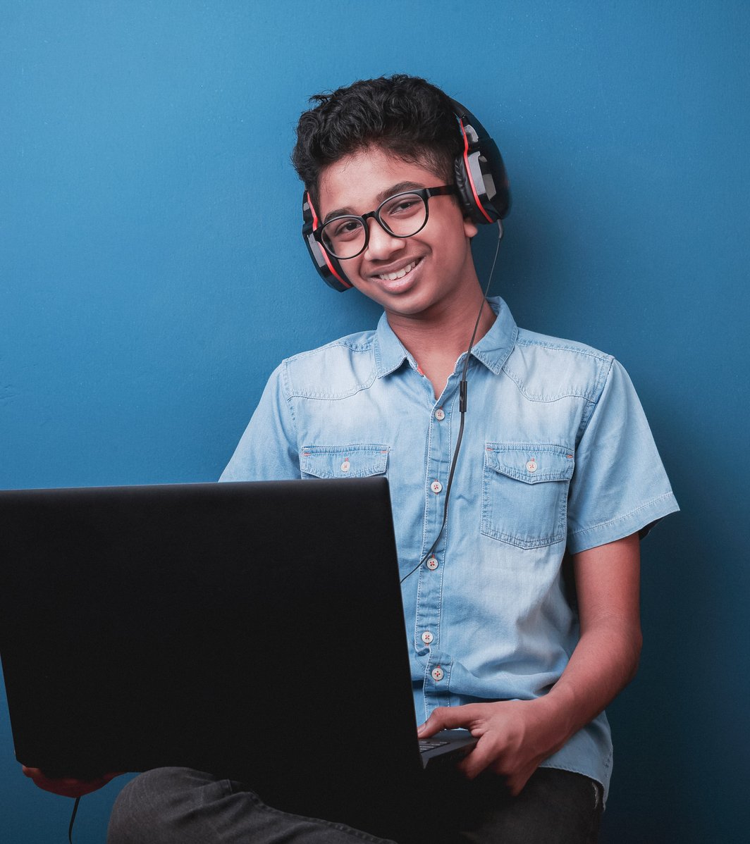 A smiling South Asian teen boy, wearing glasses and headphones, holds an open black laptop, looking directly at the camera against a plain blue background. He appears happy and engaged.