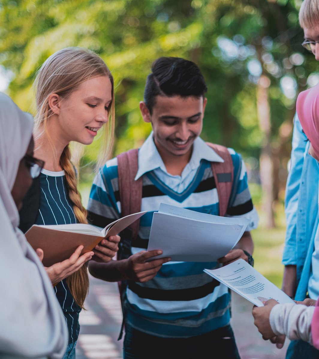 Five teenagers are gathered outdoors, smiling and looking at books and papers, engaged in a discussion or collaborative learning. The bright outdoor setting with green foliage suggests a positive and engaging environment.