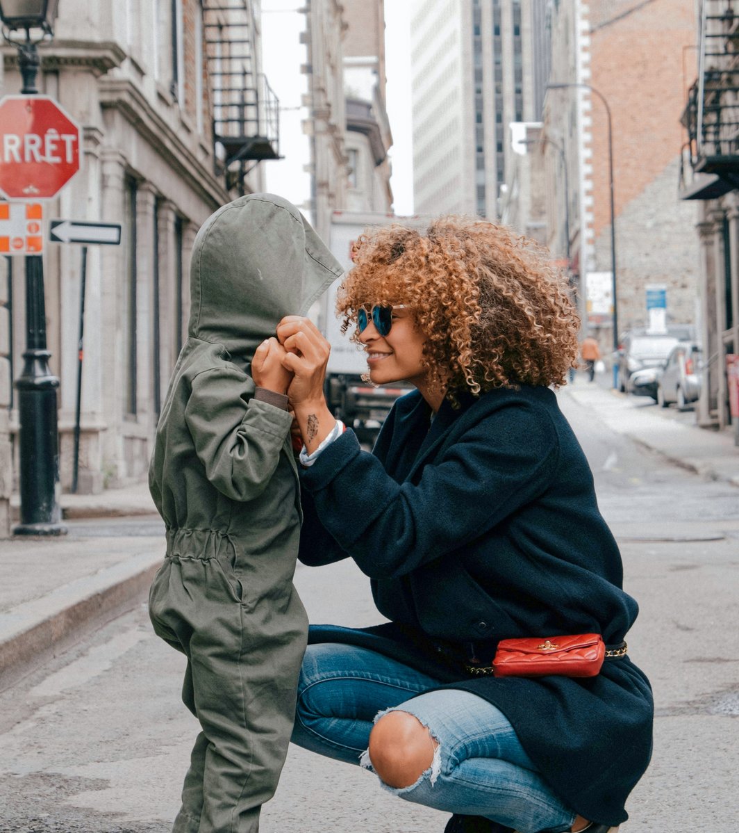 An adult with curly hair and sunglasses crouches down to hold the hands of a young child in a hooded jumpsuit on an urban street. The adult is smiling warmly at the child, conveying a sense of connection and care.