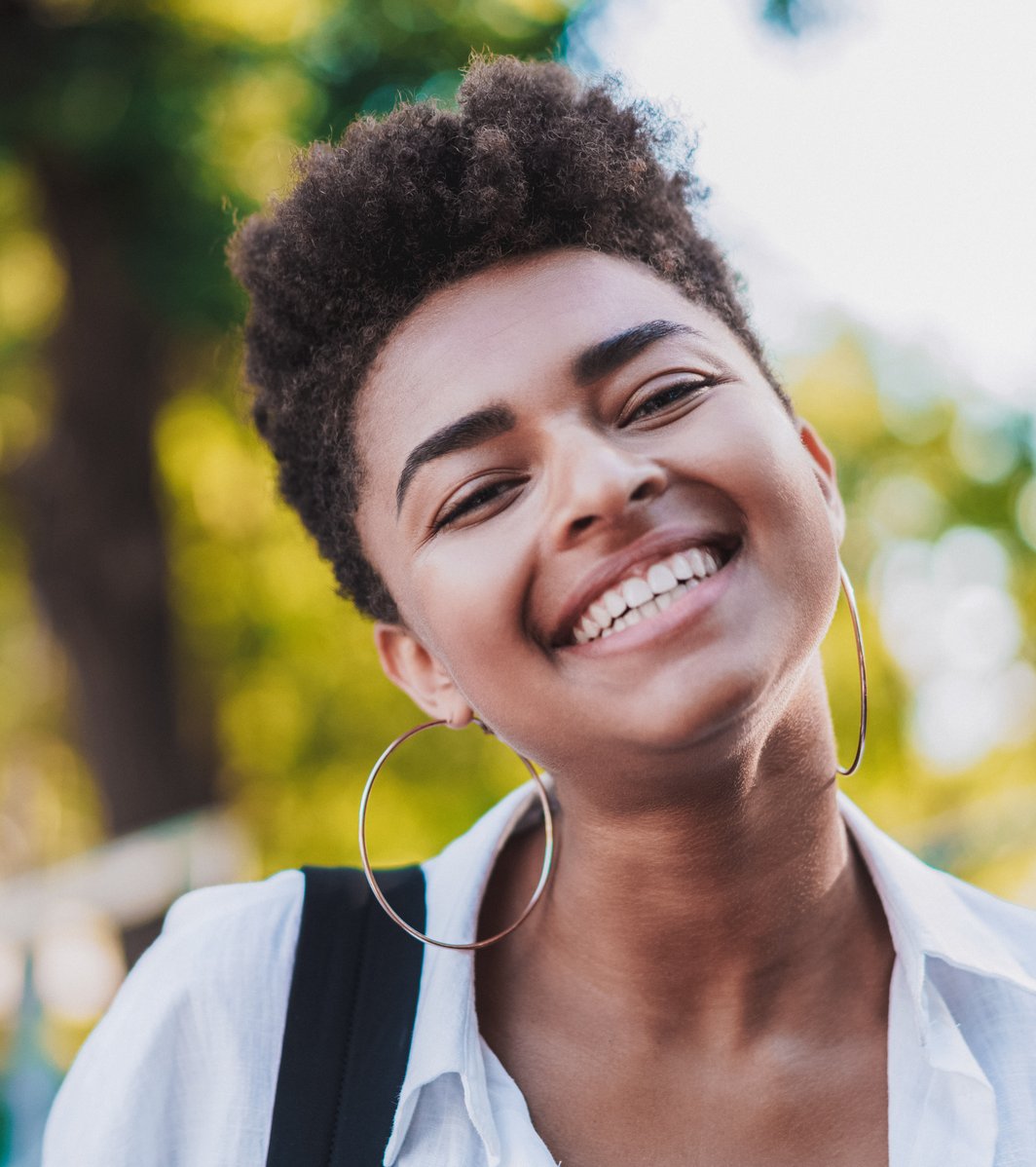 A happy teenager with short curly hair and large hoop earrings smiles widely at the camera, with a blurred green outdoor background, conveying a positive and welcoming mood.