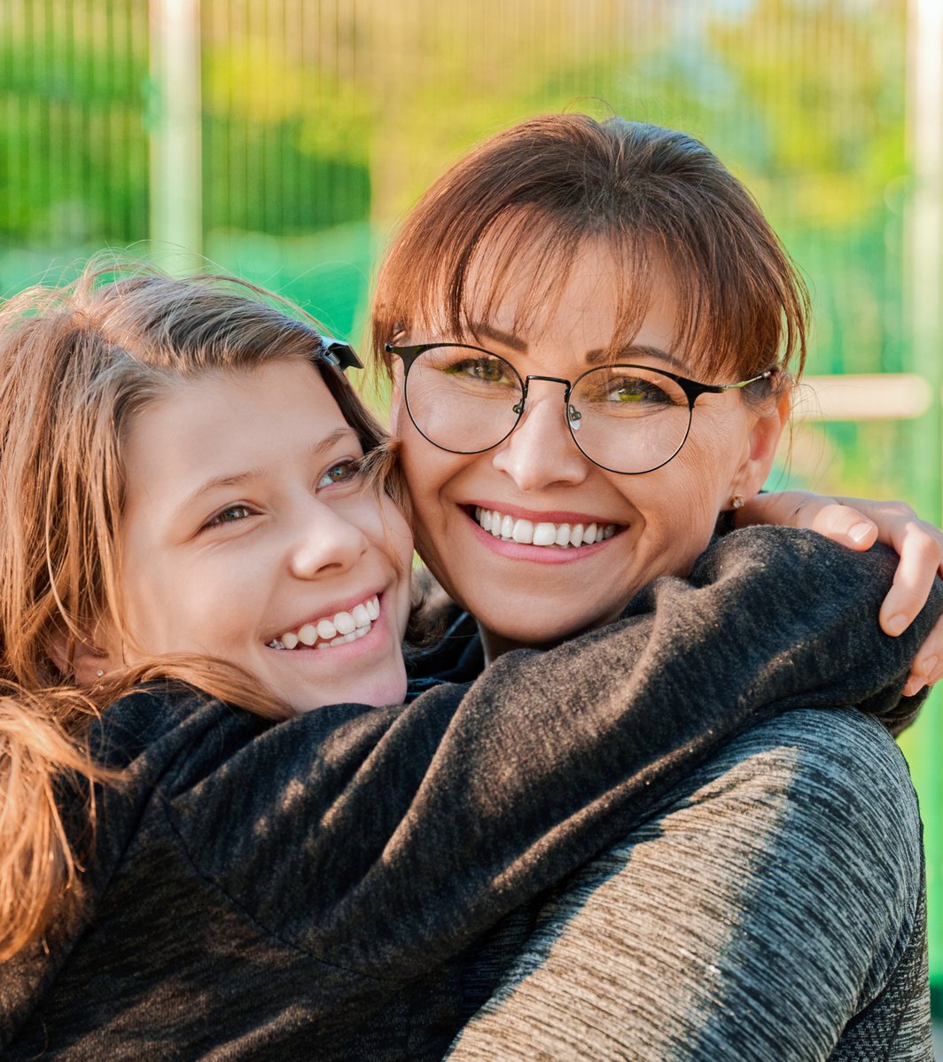 An adult woman wearing glasses is embracing a teen girl outdoors, with both smiling warmly. The background is a blurred green field.