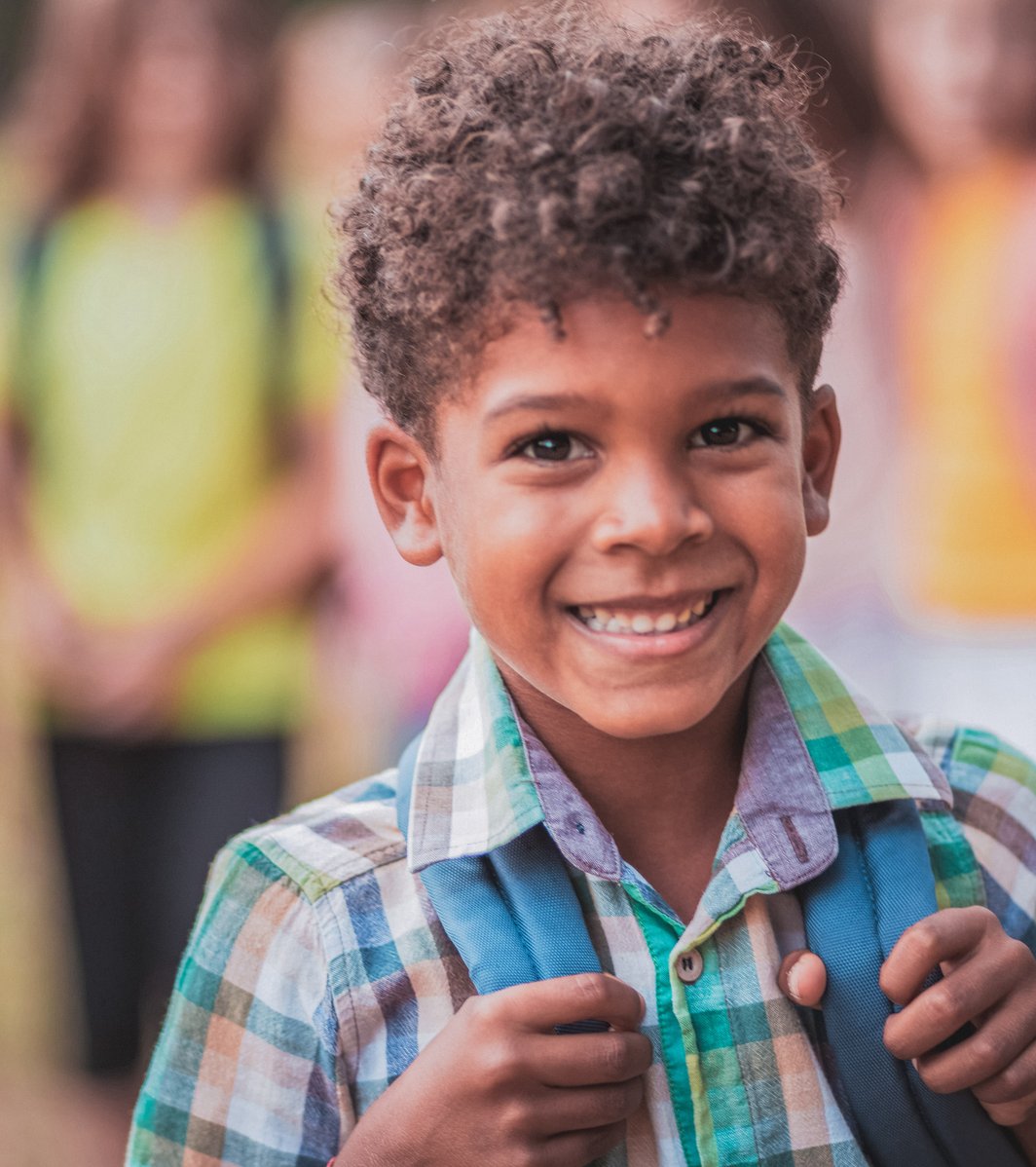A cheerful young boy with curly hair smiles brightly at the camera, wearing a plaid shirt and a blue backpack. Several other blurred children are visible in the background, suggesting an outdoor school or play setting.