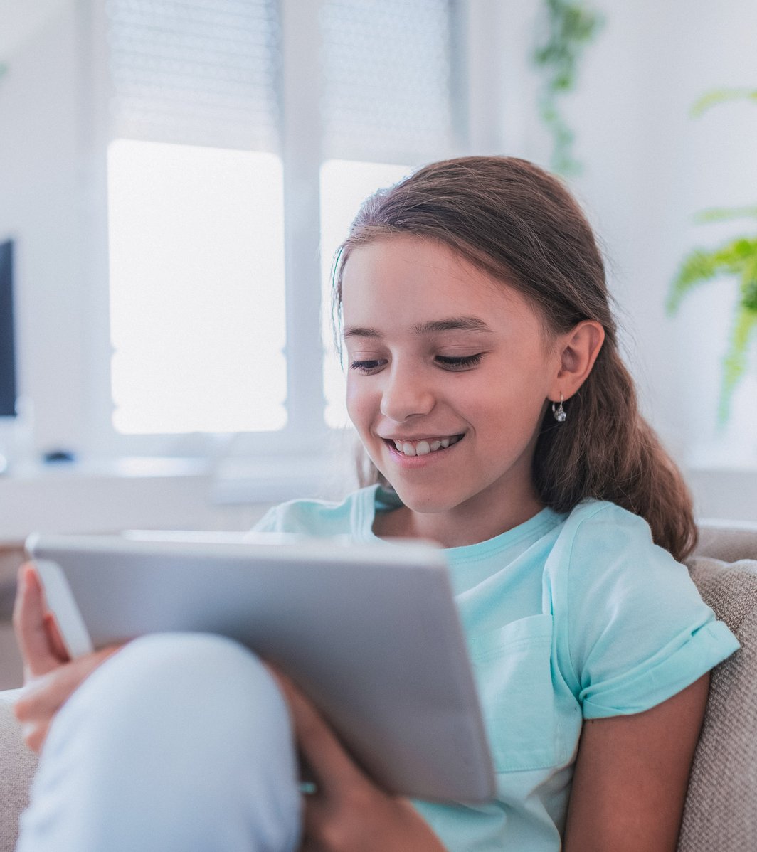 A smiling young girl sits comfortably on a light-colored couch, focused on a tablet she holds, in a bright indoor setting.
