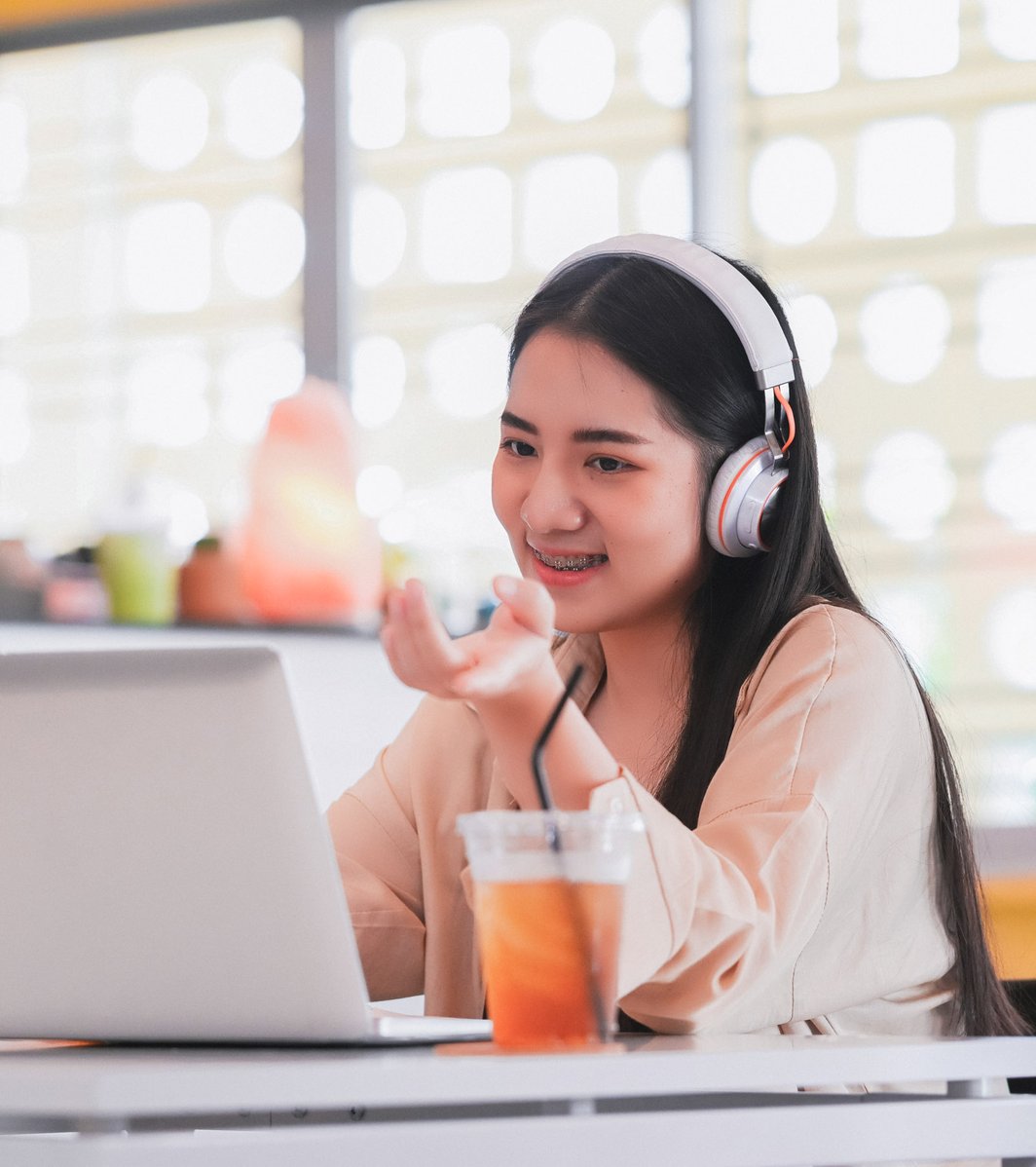 A teen wearing headphones smiles and gestures while engaged in a video call on a laptop. They are seated at a table with a drink, in a bright, modern indoor setting.