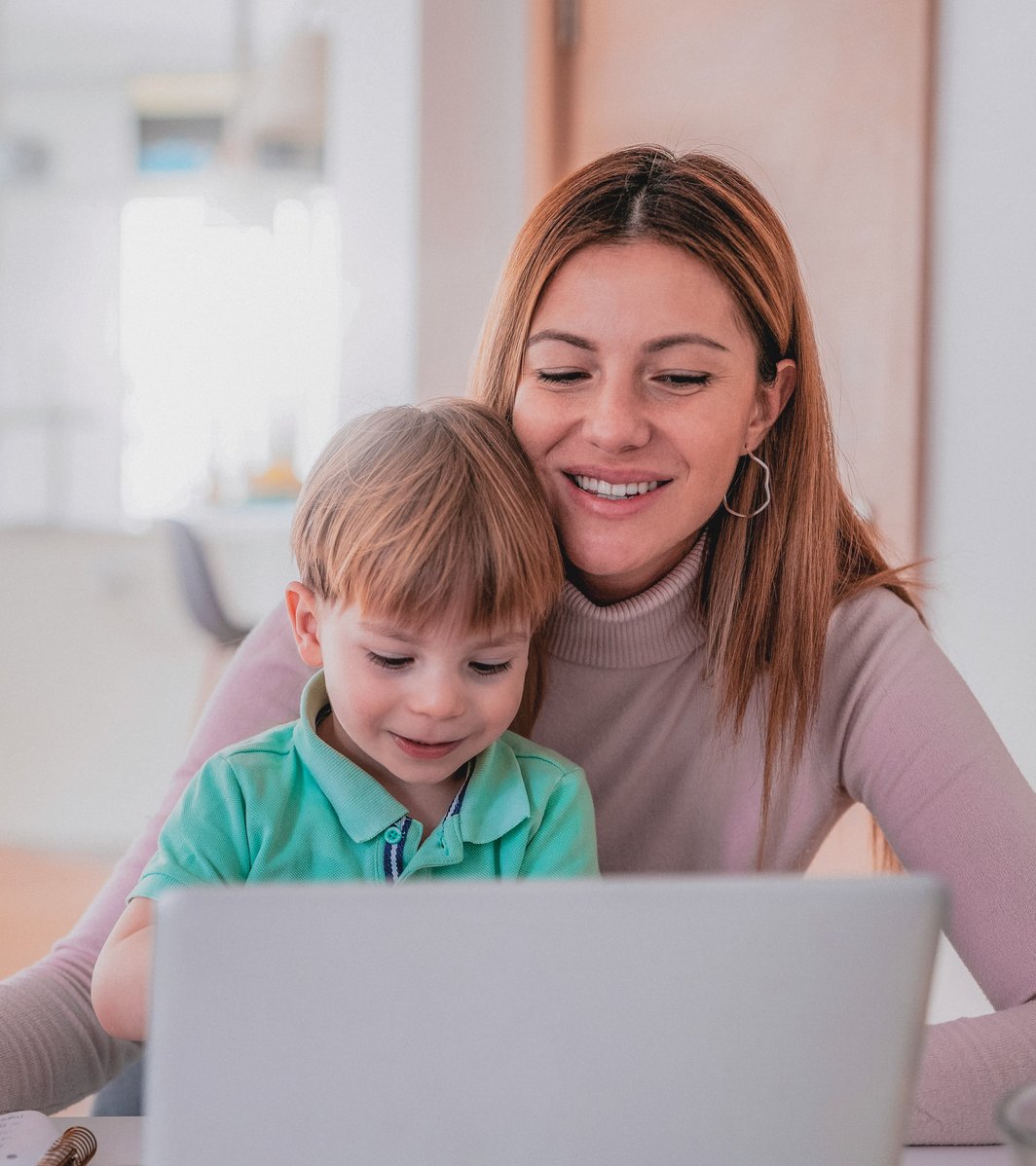 An adult woman and a young child are smiling and looking at a laptop together at a table. The woman holds a pen and a notebook, suggesting an interactive or learning moment in a bright, indoor setting.