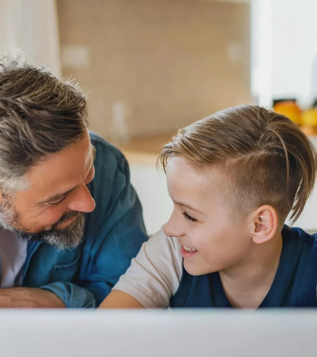 Dad and son in the kitchen smiling at each other with computer in front of them