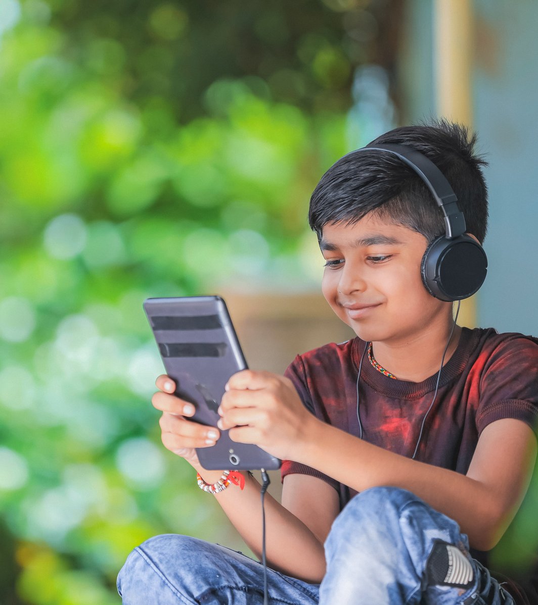 A young boy wearing black headphones is smiling while looking at a digital tablet or large smartphone. He is seated outdoors, with a blurred green leafy background, appearing calm and engaged with his device.
