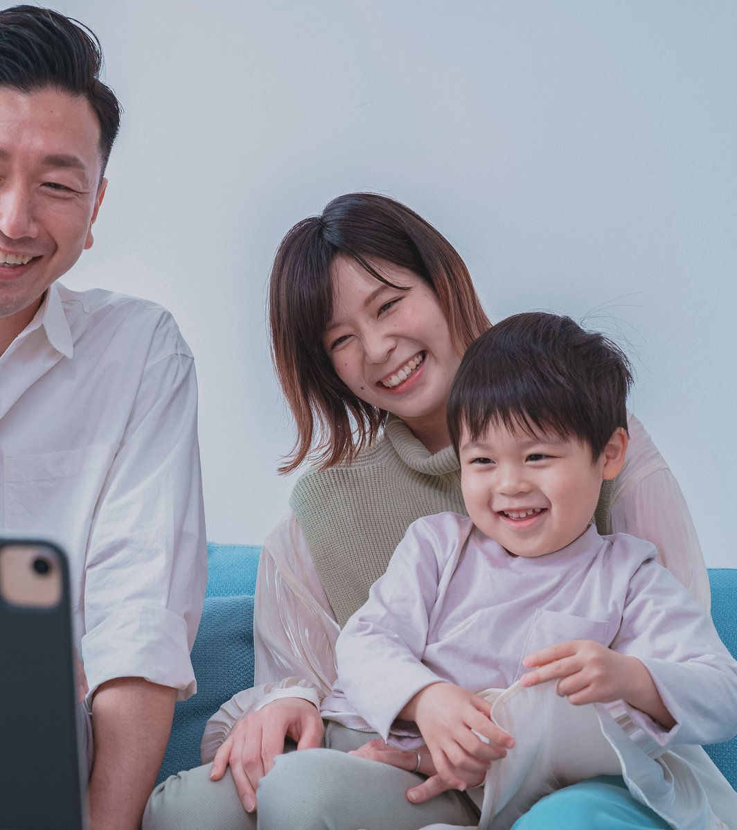 An adult man, an adult woman, and a young child are smiling happily while looking at a tablet together on a couch. They appear to be engaged in a telehealth session or video call.