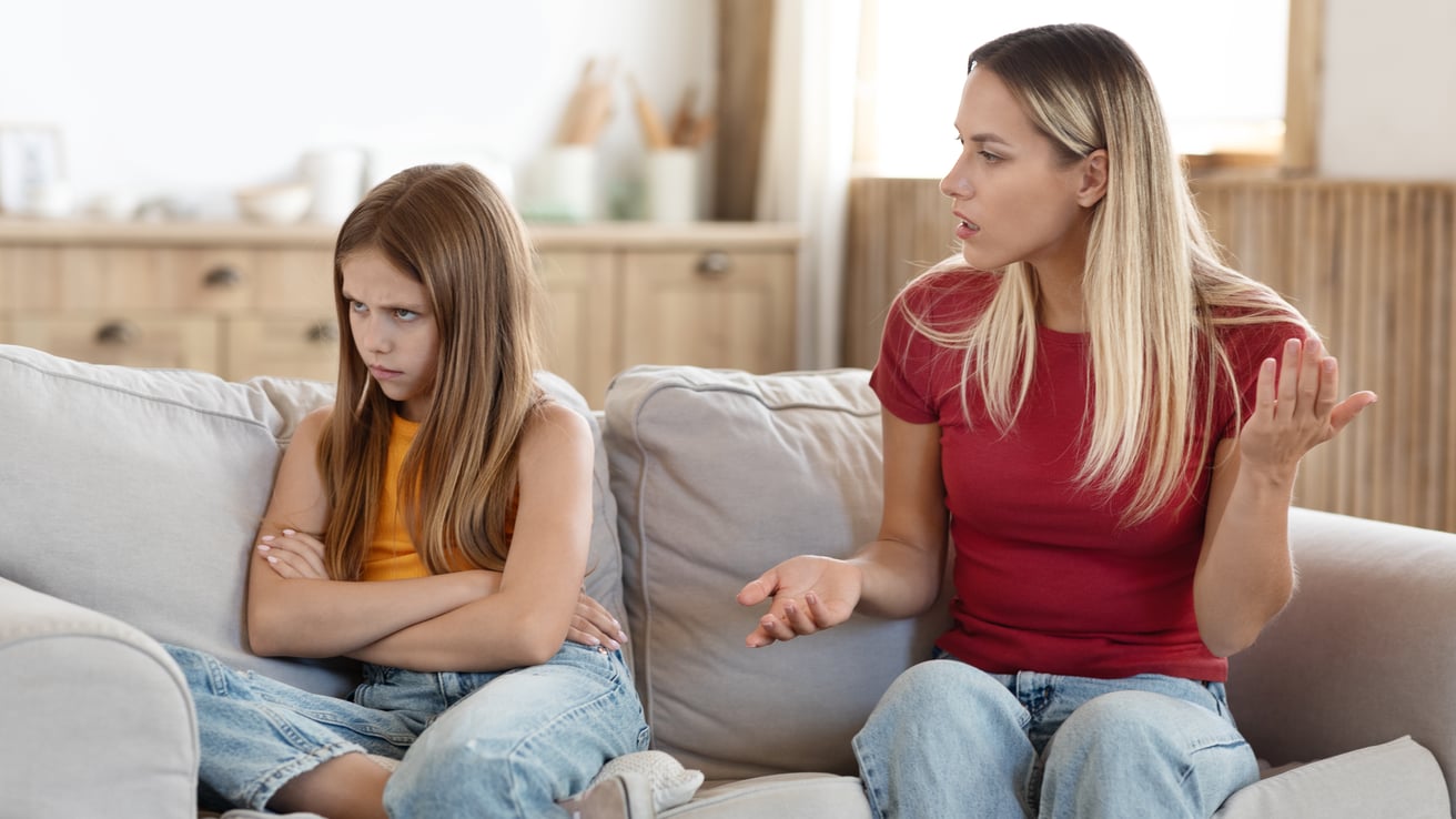 Angry teenager sits on the couch turned away from her mother who is trying to talk to her
