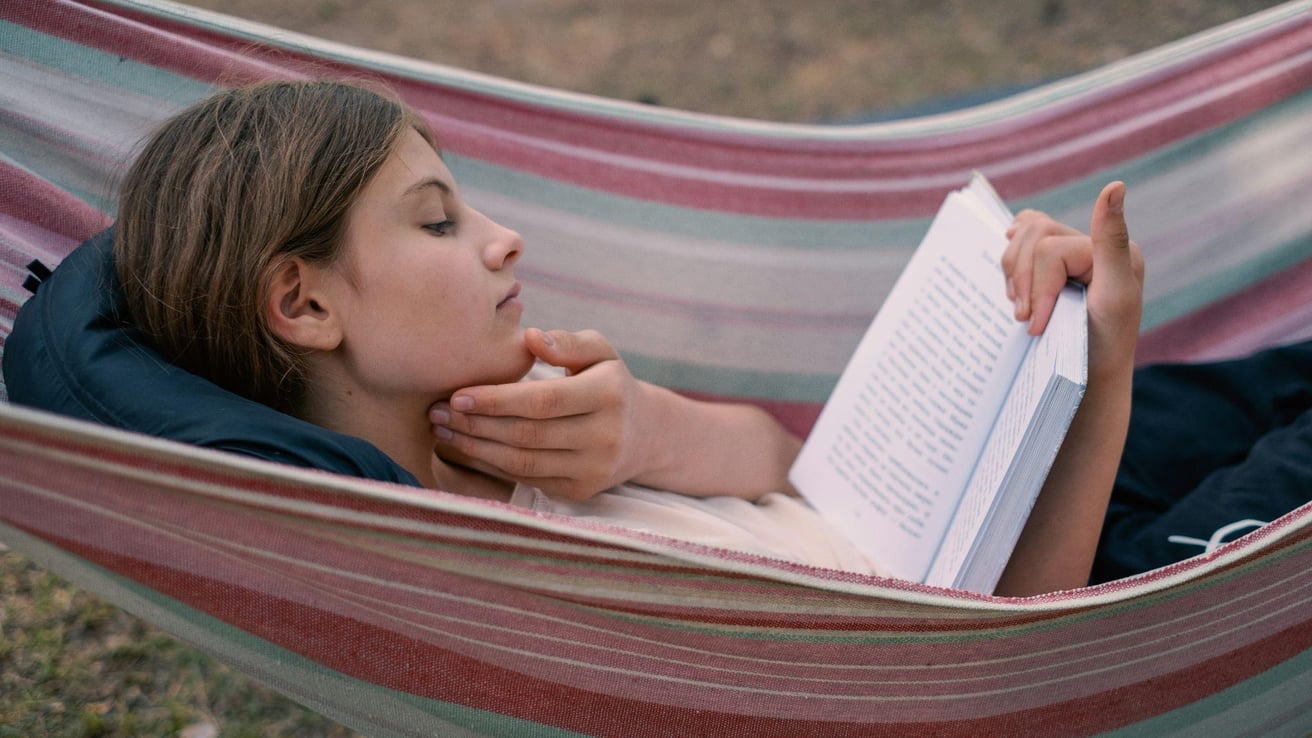 A teen lies comfortably in a striped outdoor hammock, thoughtfully reading an open book in a peaceful setting.