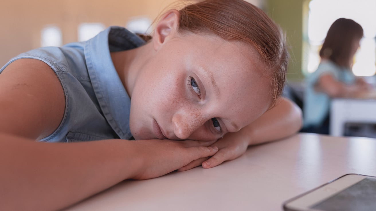 A young girl with reddish-brown hair rests her head on her arms on a table, looking directly at the viewer with a subdued and thoughtful expression. She is in an indoor setting, possibly a classroom, with a blurred figure seated at a desk in the background.