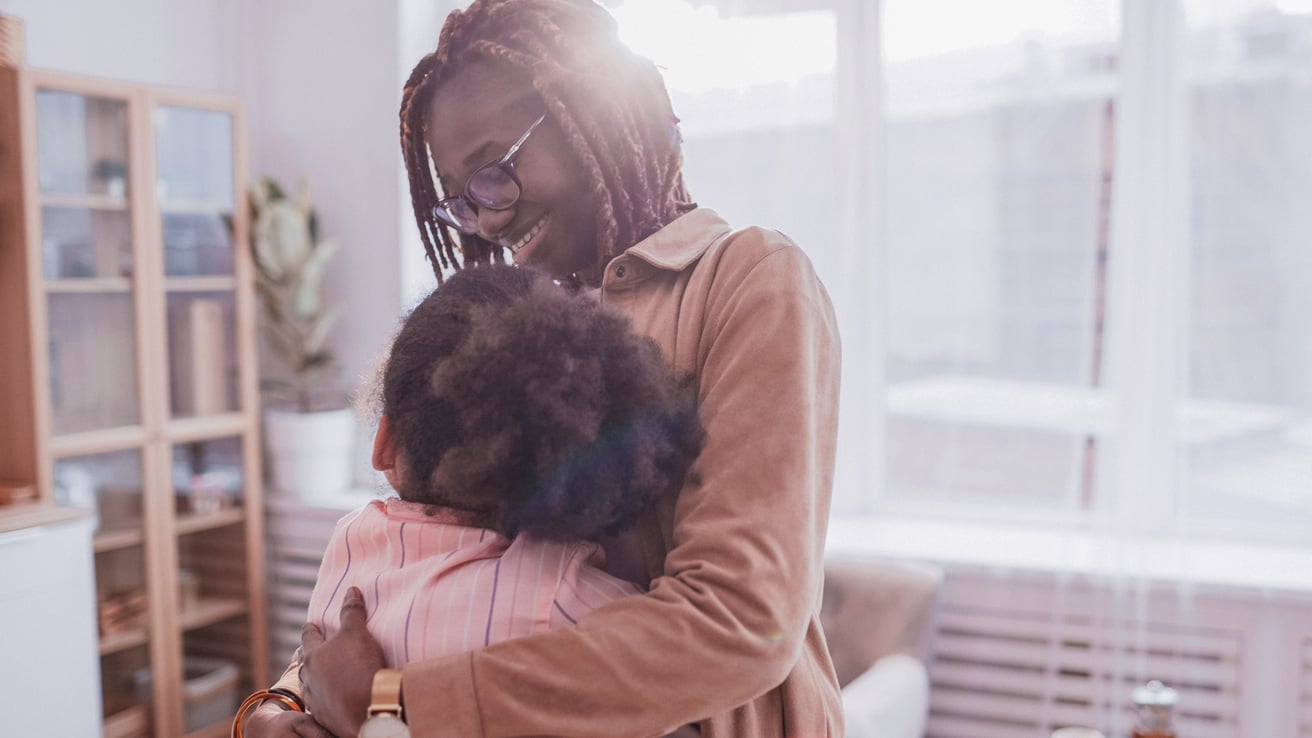 An adult with dark braids and eyeglasses is embracing a child in a brightly lit indoor space. The adult is smiling warmly, conveying a sense of comfort and affection.