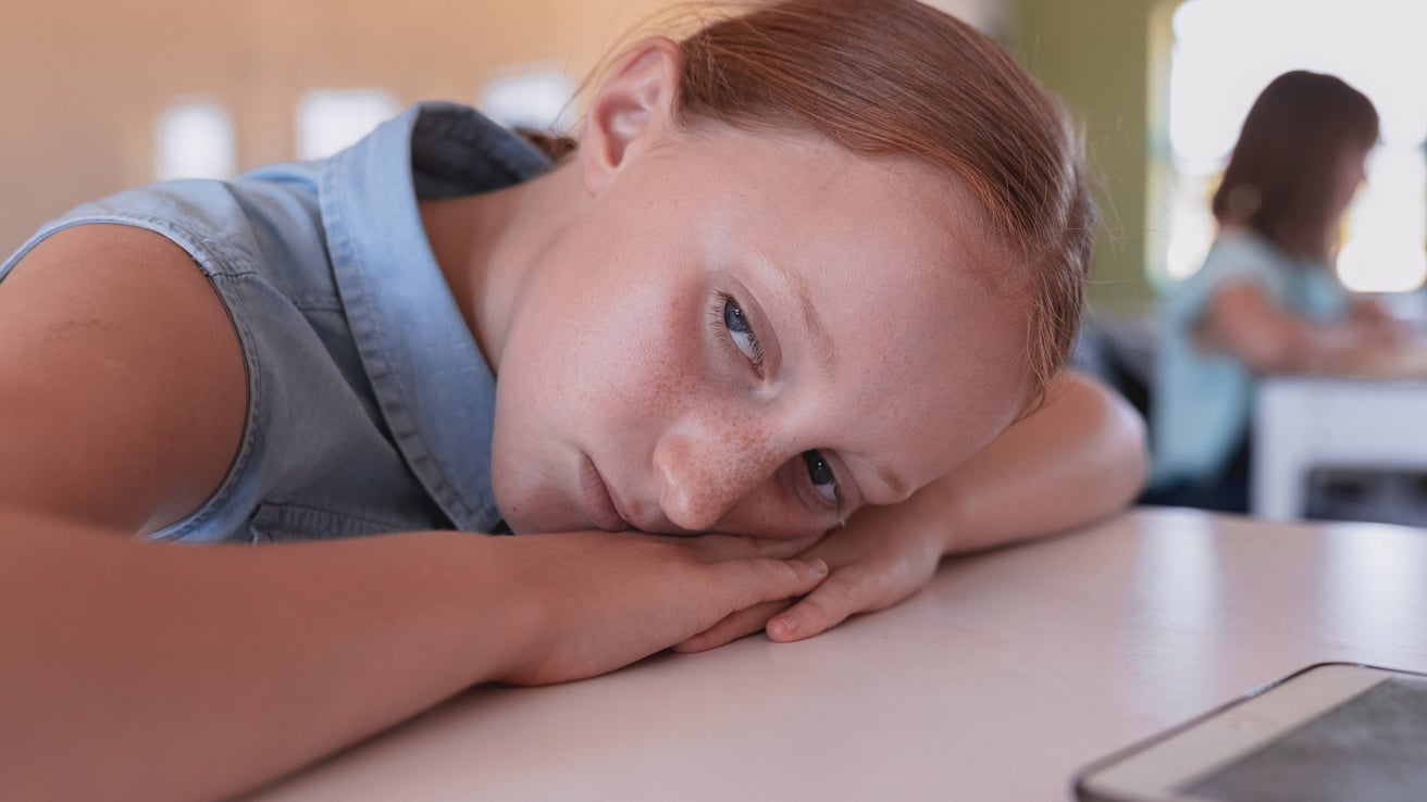 A young girl with reddish-brown hair rests her head on her arms on a table, looking directly at the viewer with a subdued and thoughtful expression. She is in an indoor setting, possibly a classroom, with a blurred figure seated at a desk in the background.
