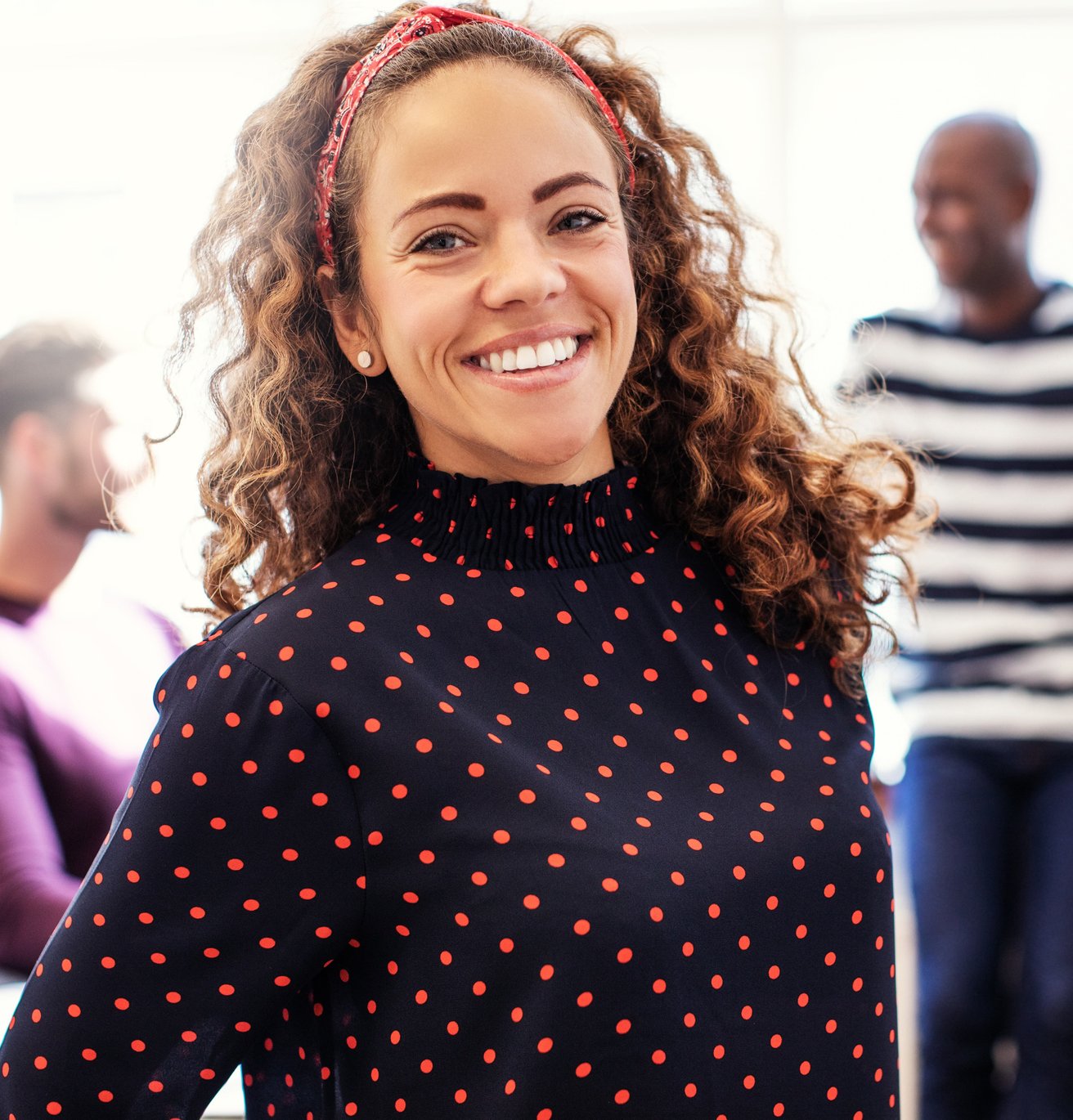 A smiling adult woman with curly hair and a red headband looks directly at the camera in a brightly lit office setting. Two blurred adult men are visible in the background, suggesting a collaborative environment.