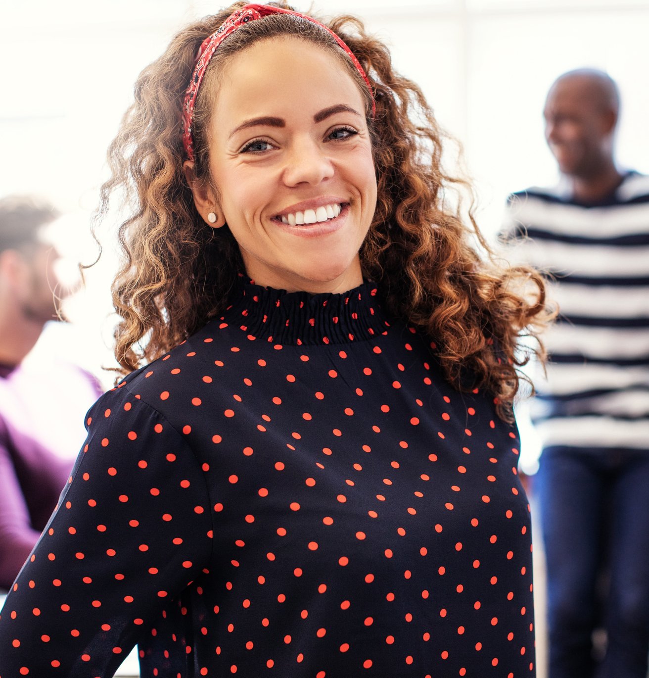 A smiling adult woman with curly hair and a red headband looks directly at the camera in a brightly lit office setting. Two blurred adult men are visible in the background, suggesting a collaborative environment.