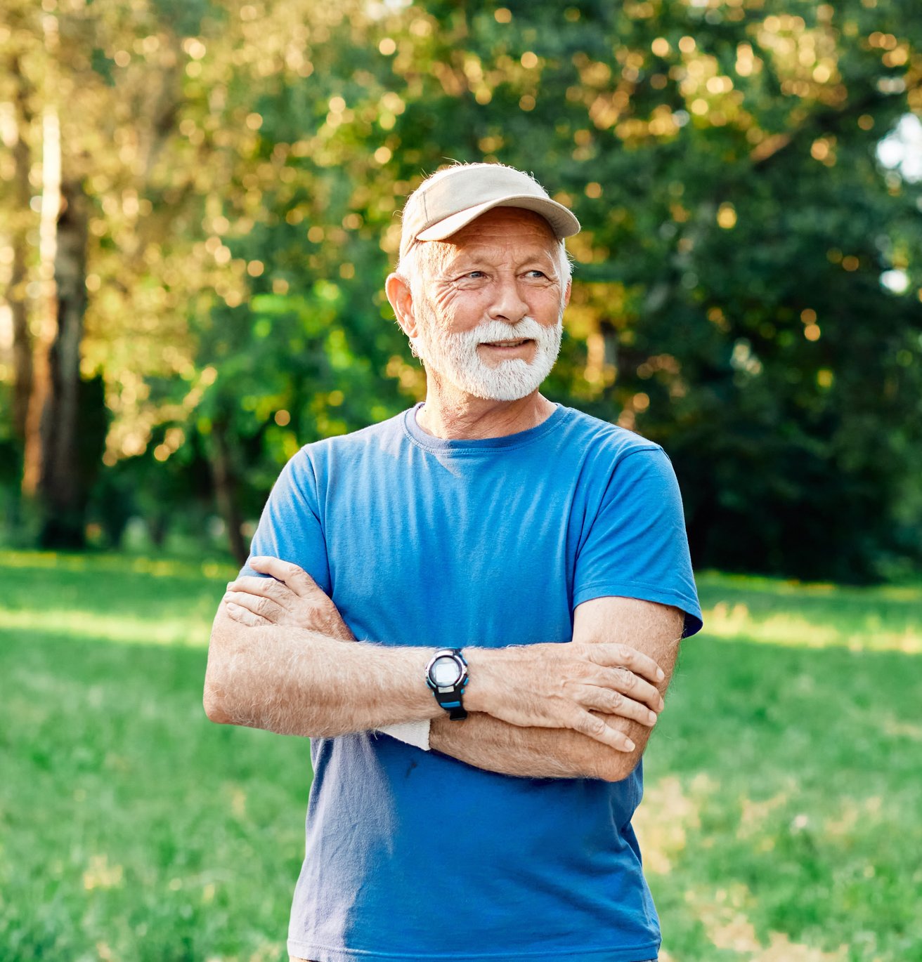 An older adult man wearing a light-colored cap and a blue t-shirt stands with his arms crossed in a sunny park. He has a pleasant expression, looking towards the side.