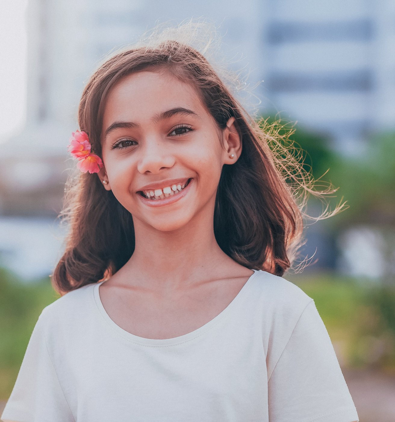 A young girl with long brown hair and a pink flower behind her ear smiles brightly. She is outdoors with a softly blurred green background.