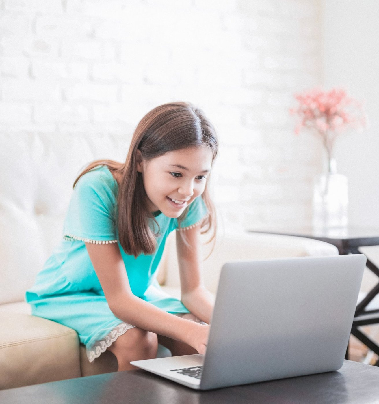 A smiling teen girl sits comfortably on a light-colored couch, looking engaged with a laptop in a bright living room setting. She appears happy and focused on the screen.