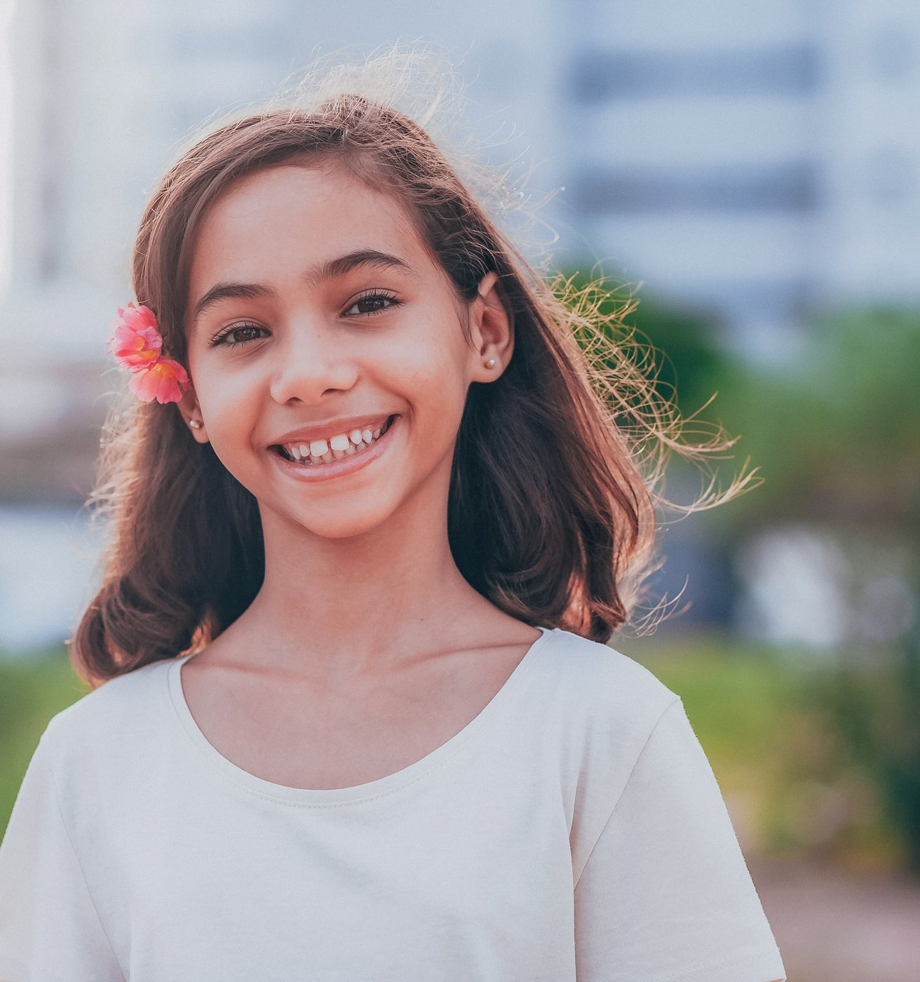 A young girl with long brown hair and a pink flower behind her ear smiles brightly. She is outdoors with a softly blurred green background.