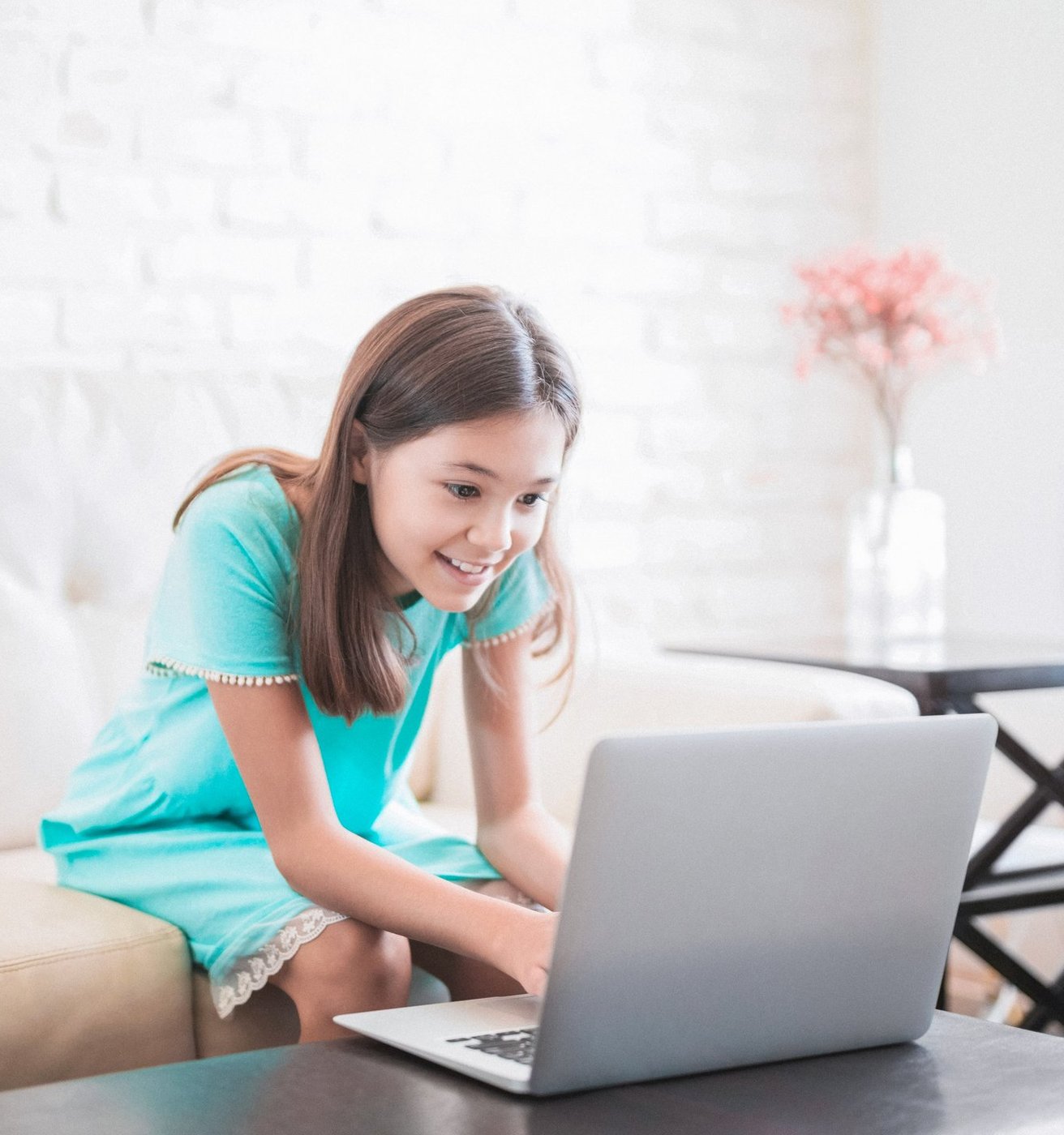 A smiling teen girl sits comfortably on a light-colored couch, looking engaged with a laptop in a bright living room setting. She appears happy and focused on the screen.