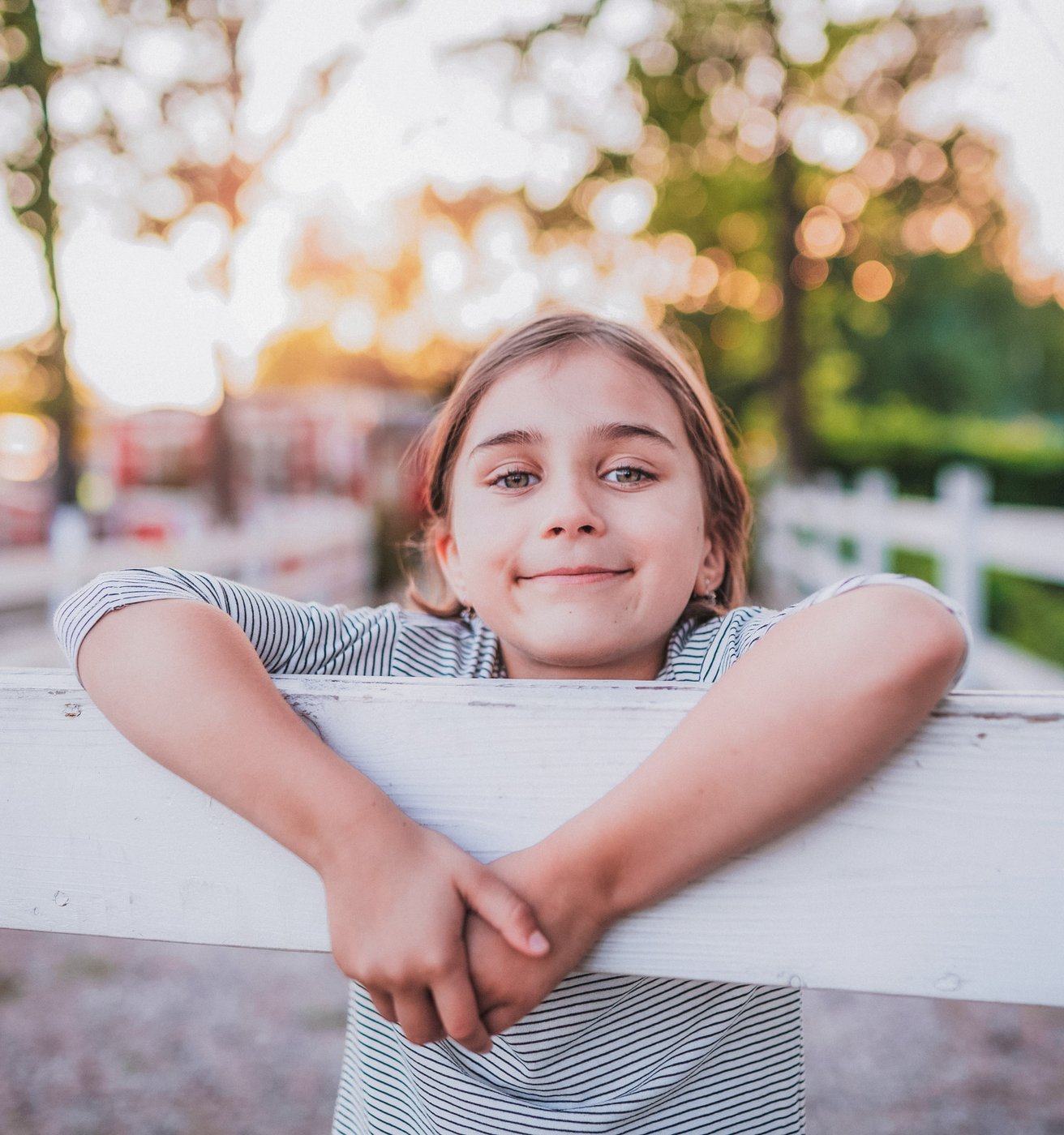 A happy young child leans on a white wooden fence outdoors, smiling gently at the viewer. The background is softly blurred with green trees and warm, bright sunlight.