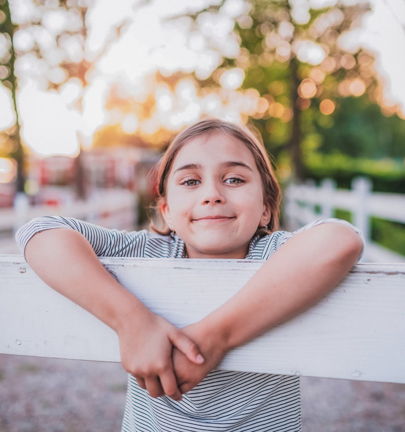 A happy young child leans on a white wooden fence outdoors, smiling gently at the viewer. The background is softly blurred with green trees and warm, bright sunlight.
