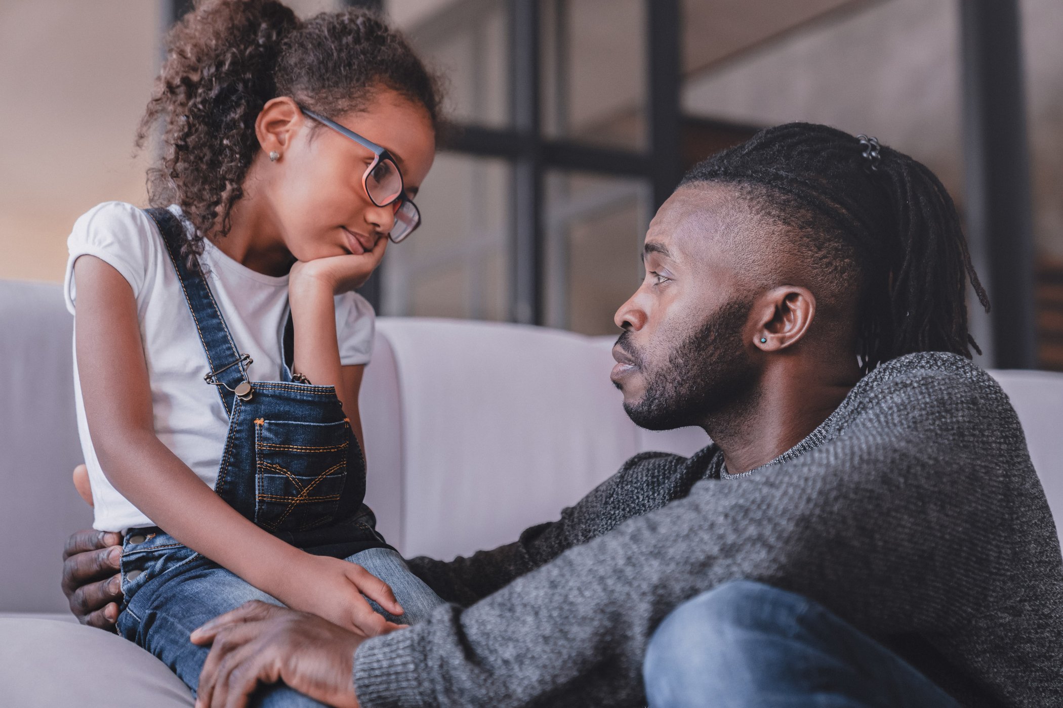 A thoughtful young girl with curly hair and glasses, wearing denim overalls, sits on a couch with an adult man who has dreadlocks and a beard. The man attentively looks at the girl, gently holding her knee, as she leans her head on her hand with a pensive expression, suggesting a serious conversation or comforting moment.