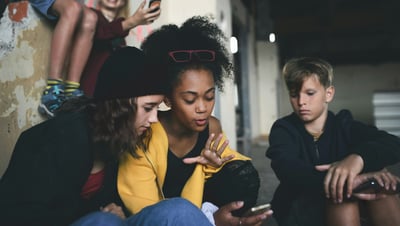 Two teenage girls are intently focused on a smartphone one of them is holding, while a teenage boy sits nearby looking down at his own phone. They are gathered in an urban, worn environment with another child visible in the background.