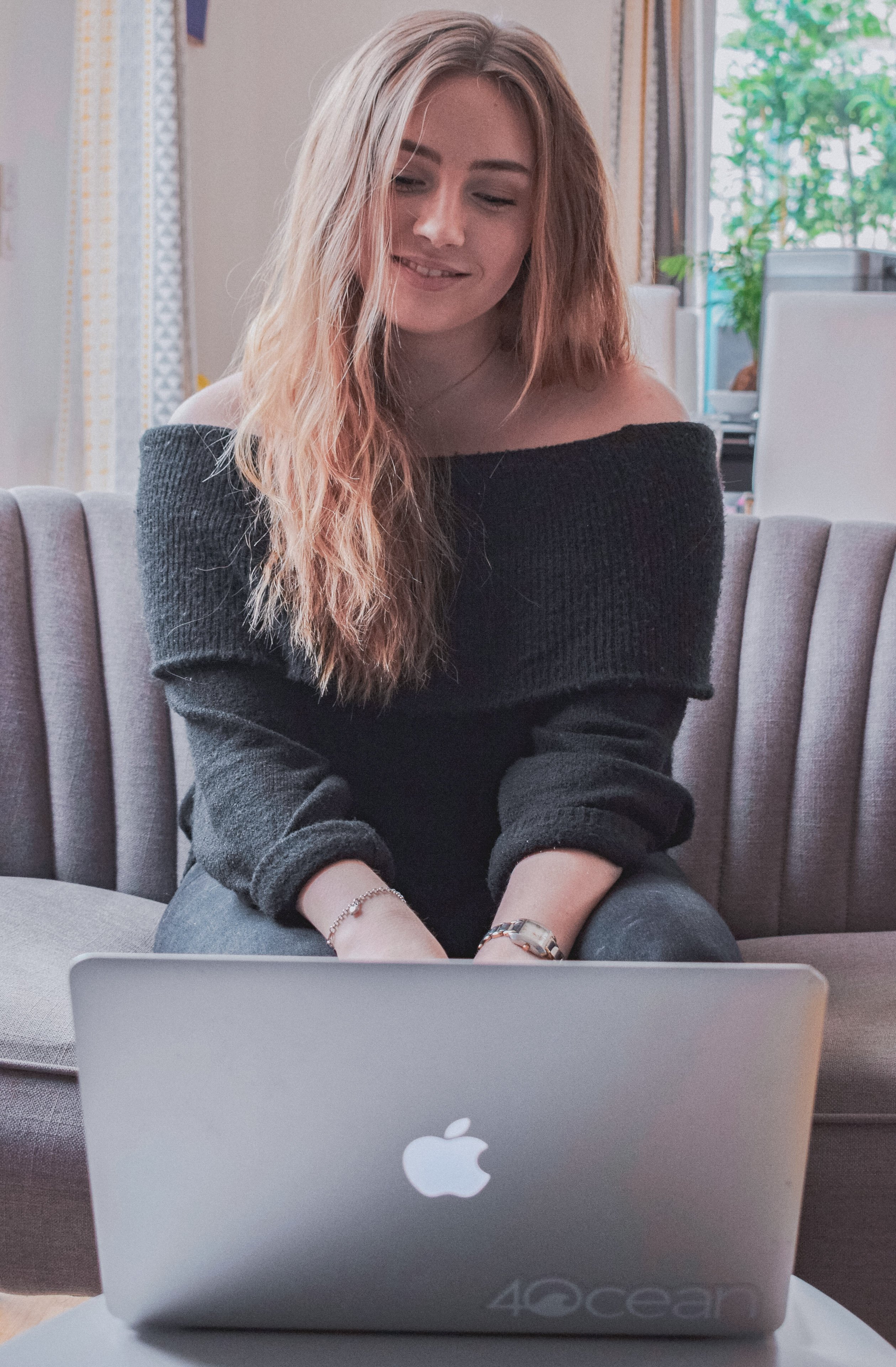 A young woman or teen sits comfortably on a grey couch, looking down at a laptop with a gentle smile, indicating an engaged online interaction in a bright indoor setting.