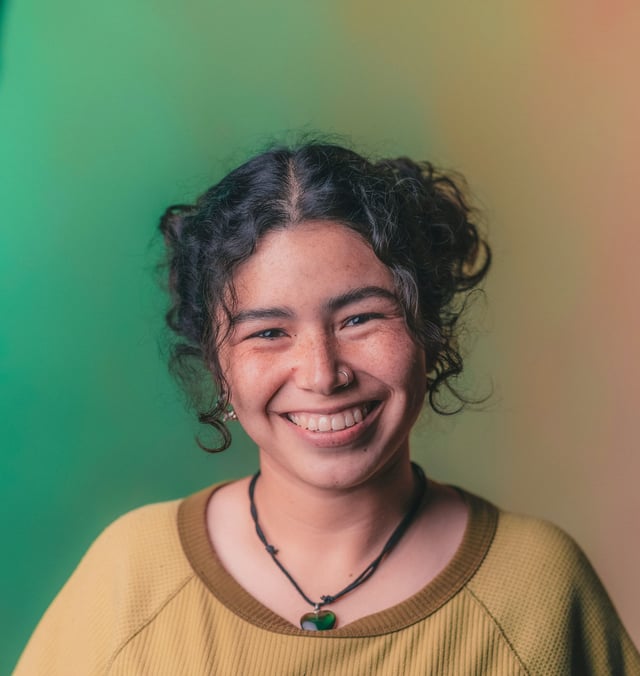 A smiling young adult with curly dark hair and freckles looks directly at the camera. They wear a yellow-green top and a necklace with a heart-shaped pendant, against a vibrant gradient background.