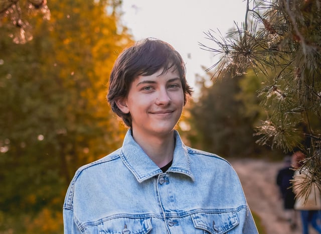 A smiling teenager with short, dark hair wears a light blue denim jacket while looking directly at the camera. They are standing outdoors in a natural setting with blurred golden-green foliage and pine needles in the background, illuminated by soft sunlight.