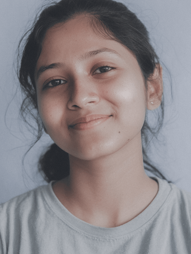 A young teenage girl with dark hair smiles gently, looking directly at the viewer in a close-up portrait against a light background. Her calm and friendly expression conveys approachability and warmth.