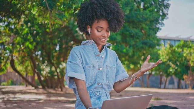 Young adult female with dark skin and a jean jacket at her laptop outside