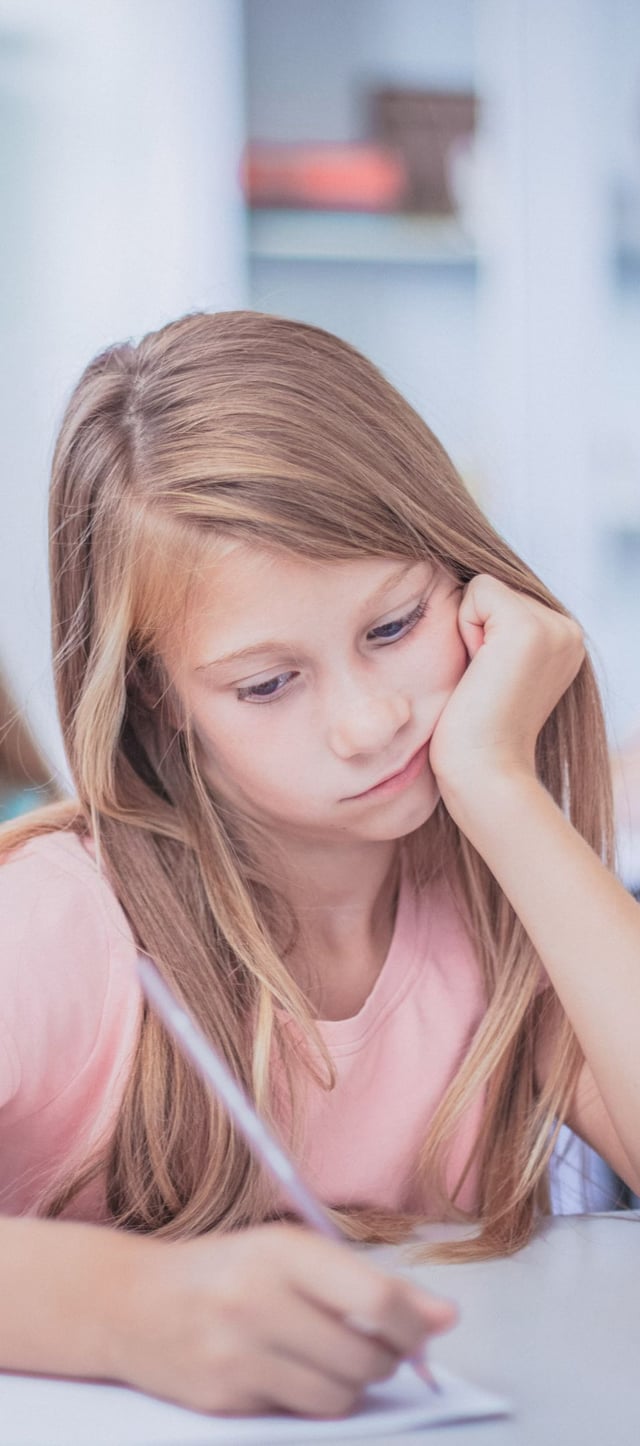 A young girl with long, light brown hair sits at a desk, concentrating while writing or drawing and resting her chin in her hand. She appears focused and thoughtful, likely in a classroom or study environment.