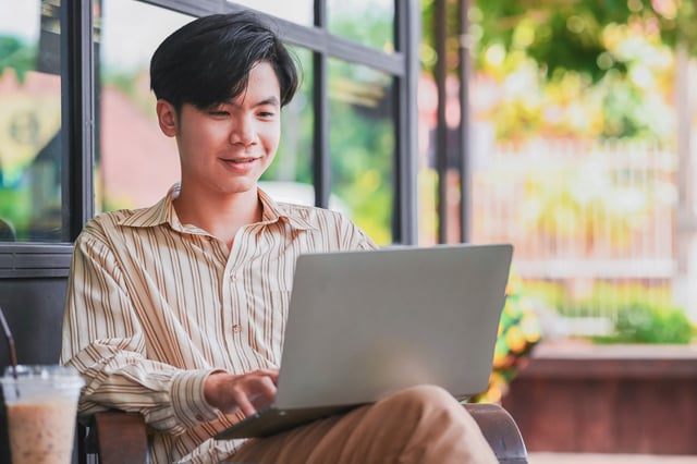 A smiling teen is comfortably sitting and using a laptop in a bright indoor space with large windows. The mood is relaxed and focused as they engage with their computer, with an iced drink visible nearby.
