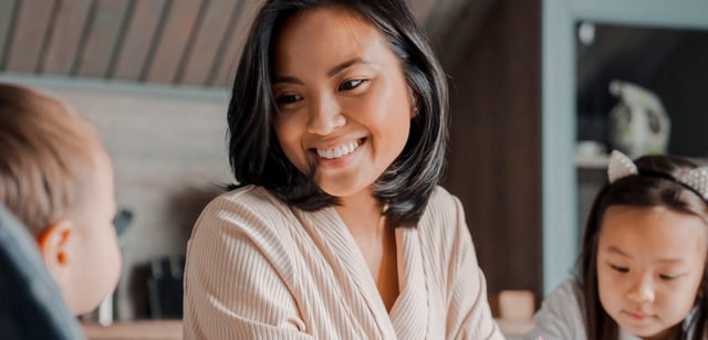 An adult woman smiles warmly while engaging with two young children indoors. A baby is partially visible on the left, and a young girl with a sparkly headband looks down on the right, creating a happy and nurturing family scene.