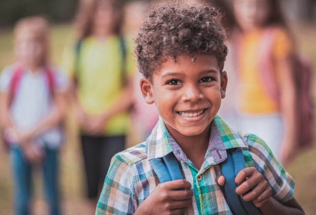 A young child, wearing a plaid shirt and a blue backpack, smiles broadly at the camera. Other children are visible, blurred, in the background outdoors, suggesting a school or play environment.