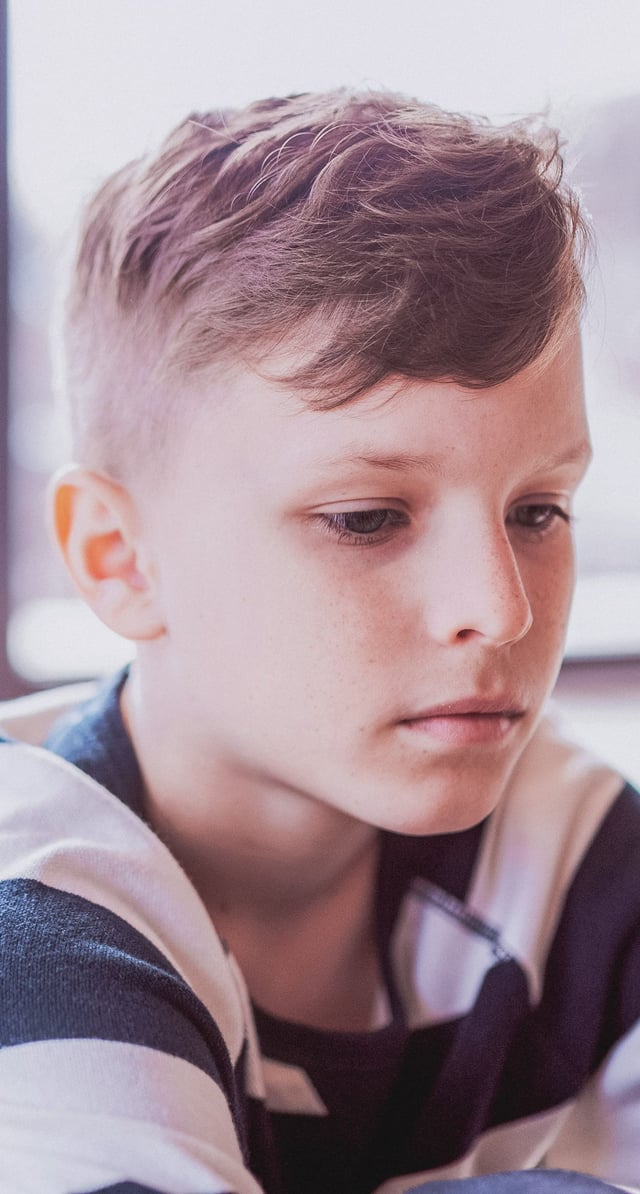 A close-up portrait of a young boy, appearing to be a pre-teen, looking down with a pensive and thoughtful expression. He is wearing a striped shirt indoors, with soft lighting on his face.