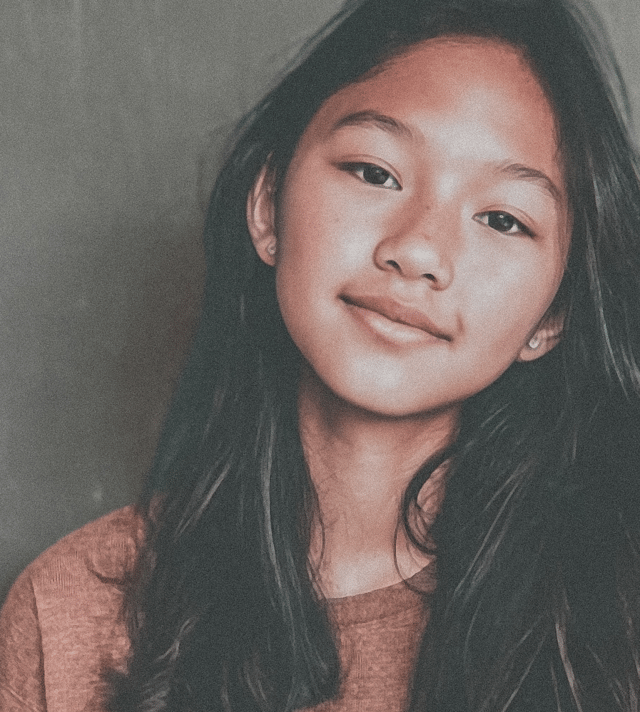 A young teen girl of East Asian ethnicity with long dark hair smiles gently, looking directly at the camera in a close-up portrait. She conveys a calm and friendly mood against a simple background.