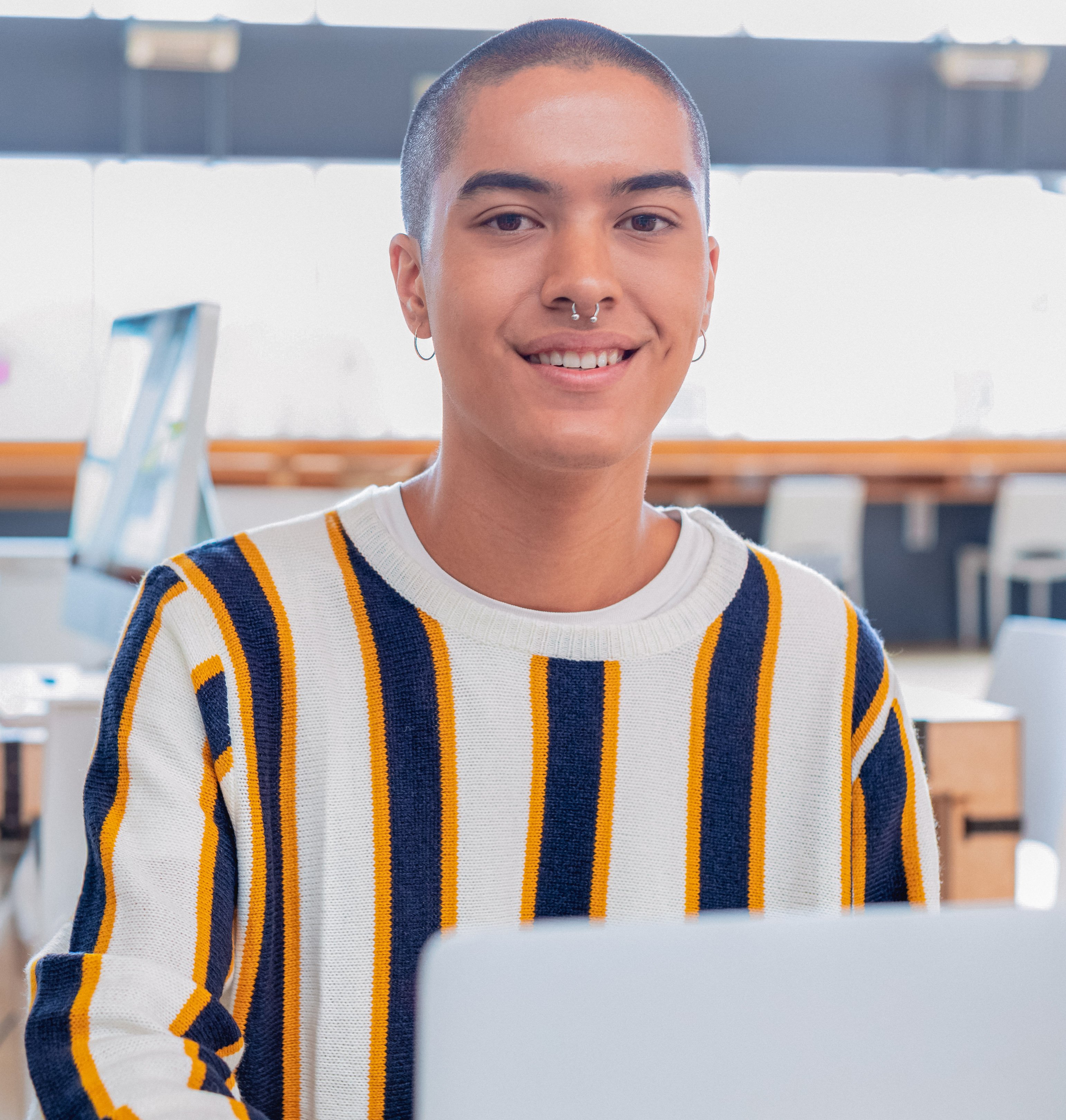 A smiling young adult with a shaved head and nose piercing looks directly at the camera while sitting at a desk with a laptop in a bright, modern indoor setting. They appear friendly and engaged.