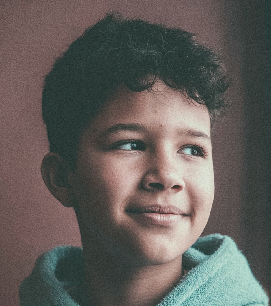 A young boy with dark curly hair looks gently to the side with a soft, pensive smile. He is wearing a light blue hooded garment, captured in a close-up portrait against a muted background.