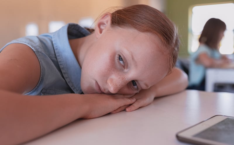 A young girl with reddish-brown hair rests her head on her arms on a table, looking directly at the viewer with a subdued and thoughtful expression. She is in an indoor setting, possibly a classroom, with a blurred figure seated at a desk in the background.