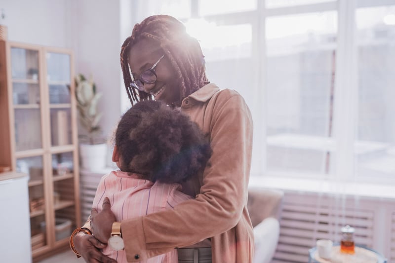 An adult with dark braids and eyeglasses is embracing a child in a brightly lit indoor space. The adult is smiling warmly, conveying a sense of comfort and affection.